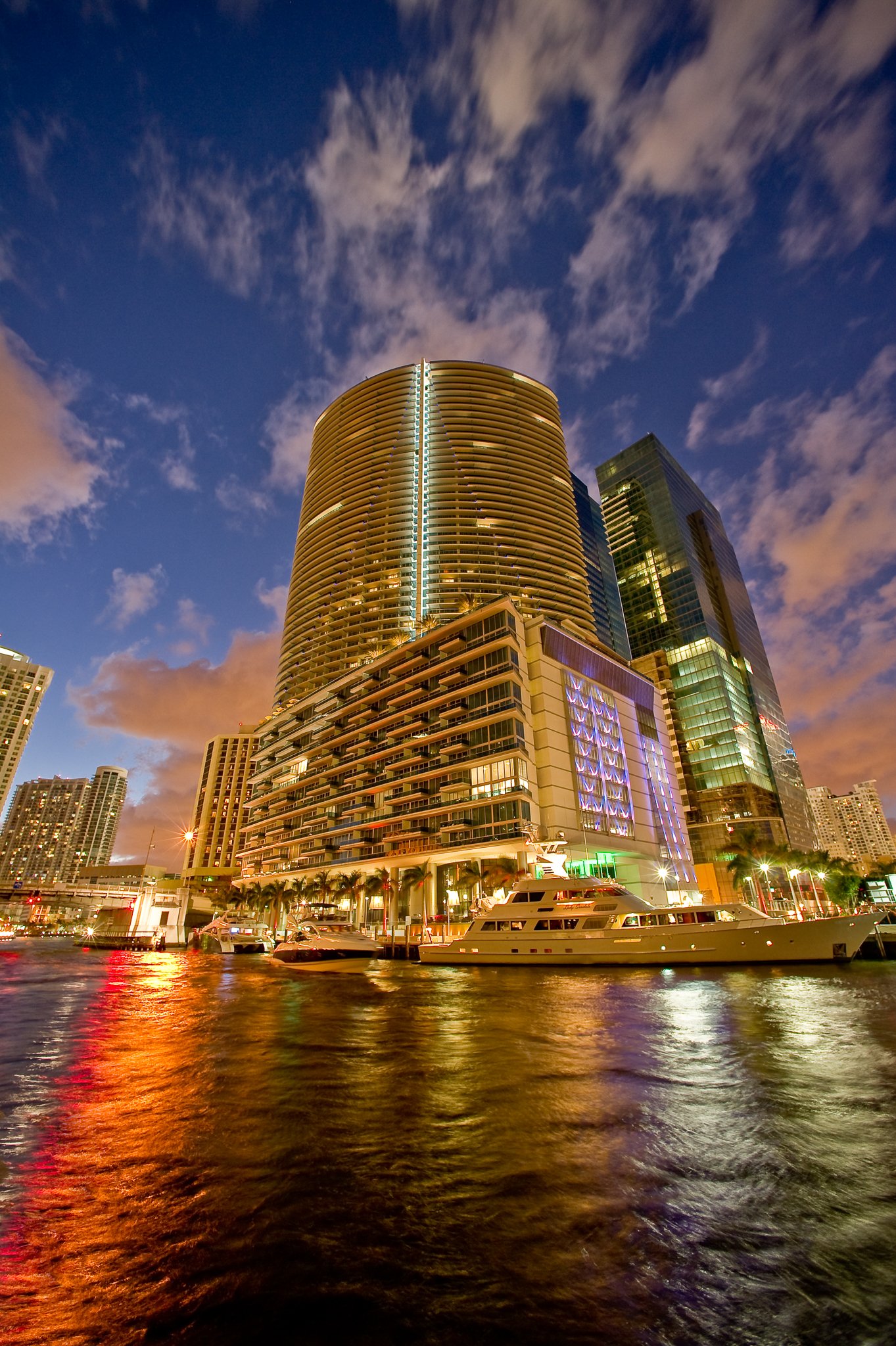 Night view of modern high-rise buildings along a waterfront, illuminated with colorful lights, with boats docked by the pier.