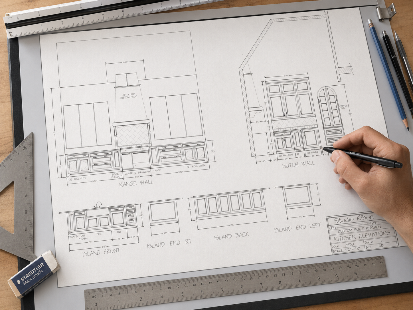 Overhead view of a large architectural kitchen elevation drawing on a drafting table, with rulers, pencils, and an eraser around the page, while a hand holding a drafting pen appears to be sketching details near the hutch wall section.