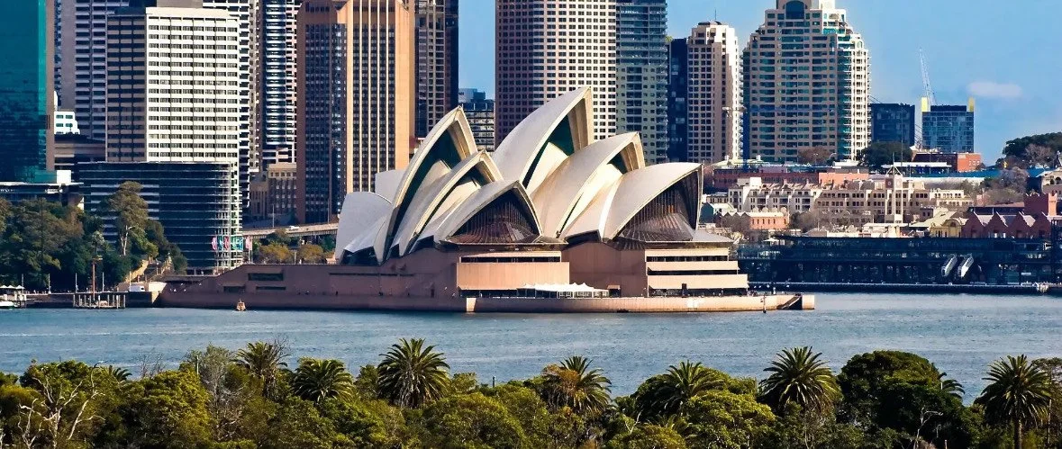 Sydney Opera House with city skyscrapers and greenery in the foreground.