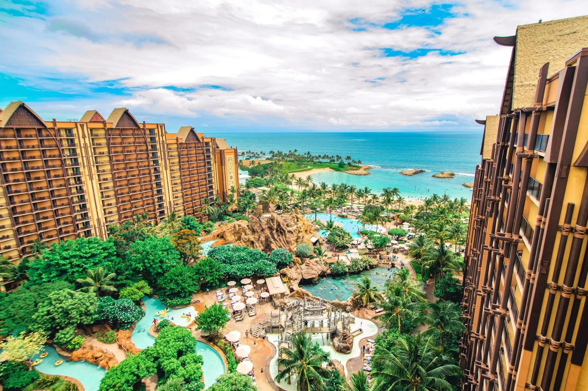 Aerial view of a tropical resort with multiple pools, lush greenery, palm trees, and beachfront with ocean in the background, taken from a high balcony or building.