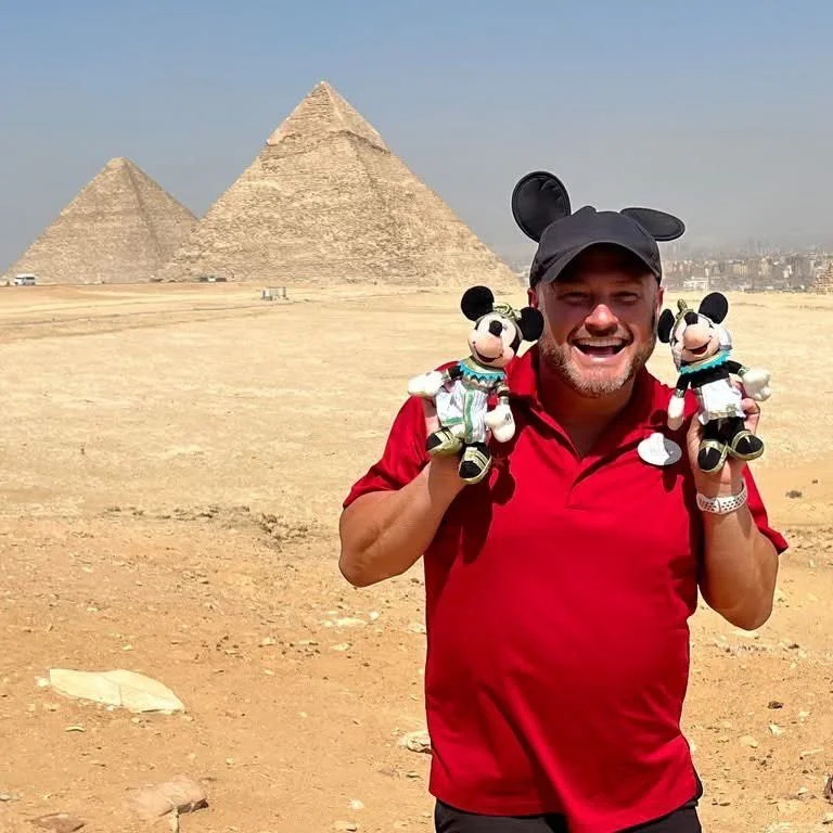 A smiling man wearing a red shirt and Mickey Mouse ears hat holding Mickey Mouse plush toys in both hands, standing in front of the pyramids of Egypt.