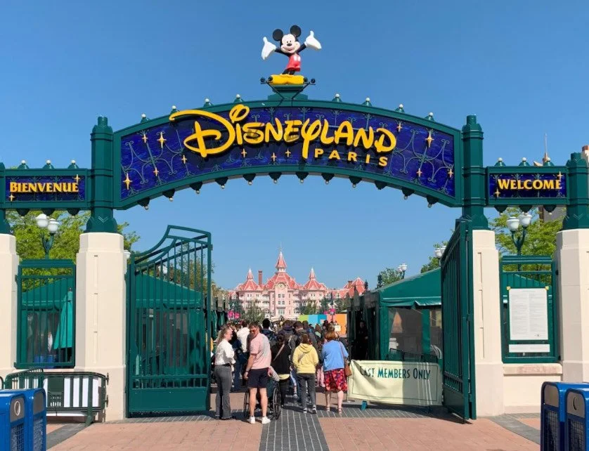 Entrance gate to Disneyland Paris with a large Mickey Mouse figure on top, colorful sign reading 'Disneyland Paris' and welcoming signs in French and English, with people entering the park and Sleeping Beauty Castle in the background.