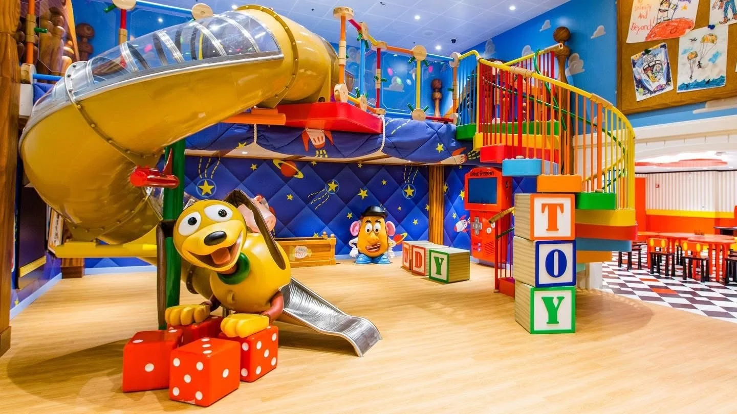 Colorful indoor children's play area with a yellow spiral slide, oversized toy blocks spelling out 'TOY', and various play structures, with cartoon character figures in the background and a blue sky with clouds painted on the wall.