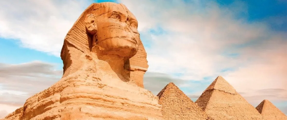 Large stone sculpture of an ancient Egyptian pharaoh's head with pyramids in the background under a cloudy sky.