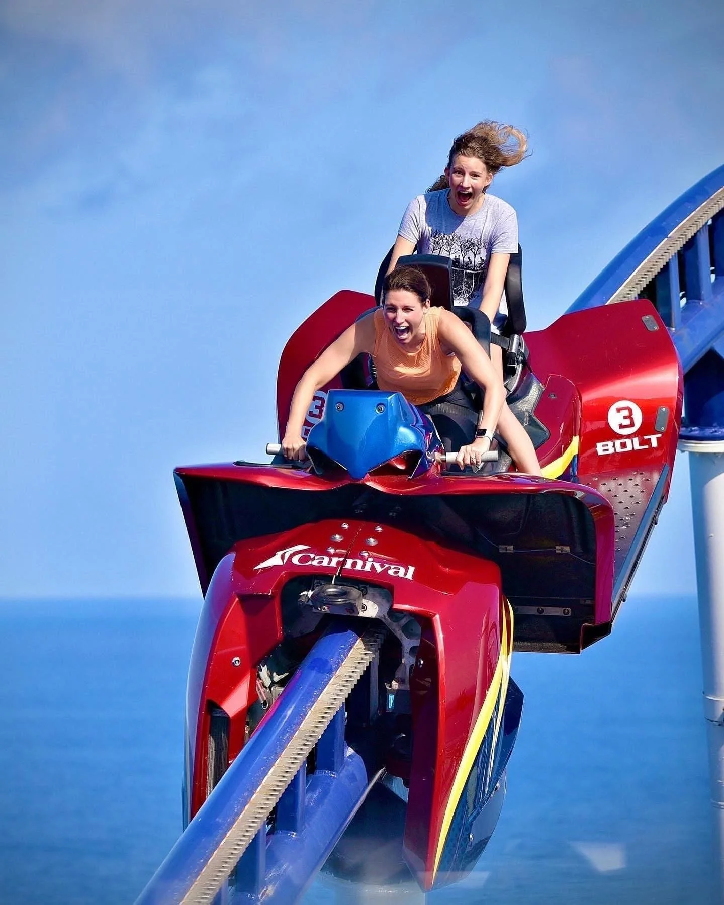 Two women are on a red roller coaster ride, with one woman in an orange shirt and the other in a gray shirt, both with expressions of excitement and joy as they descend.