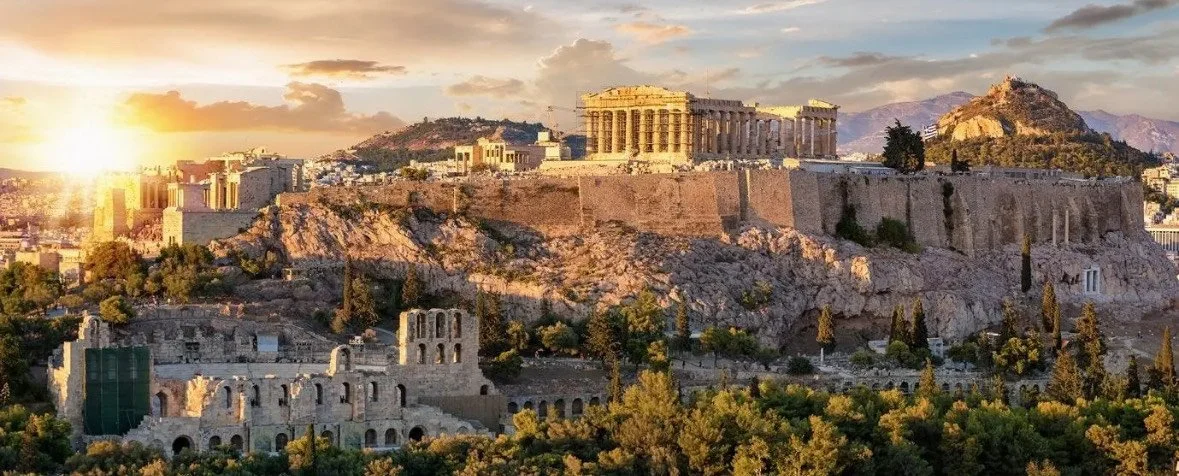 Sunset over the Acropolis in Athens, Greece, with ancient ruins and the Parthenon visible on a rocky hill, surrounded by trees and cityscape.