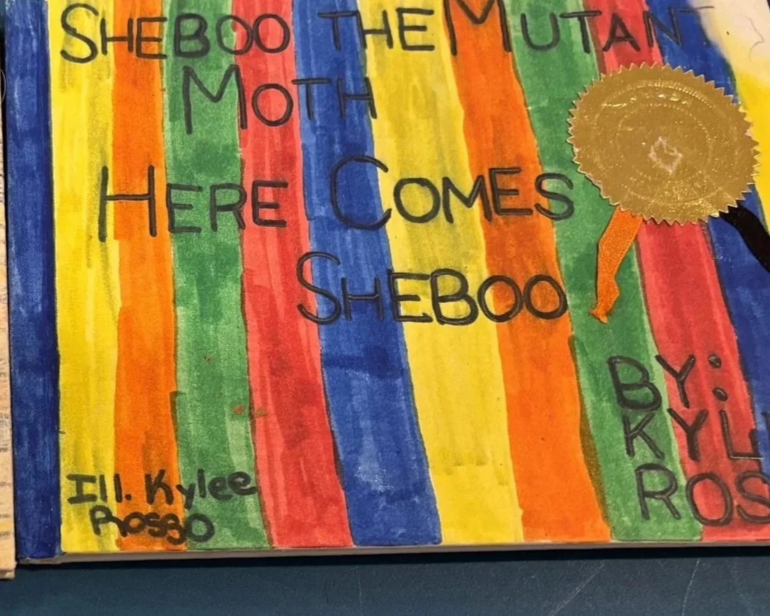 Handmade poster with rainbow-colored vertical stripes, blue, green, yellow, orange, and red, containing handwritten text in black marker. The text reads, 'SHEBOO THE MYUTAN MOT HERE COMES SHEBOO.' There is a gold seal or sticker on the right side of the poster with a black handle attached. The bottom left corner has additional smaller handwritten text