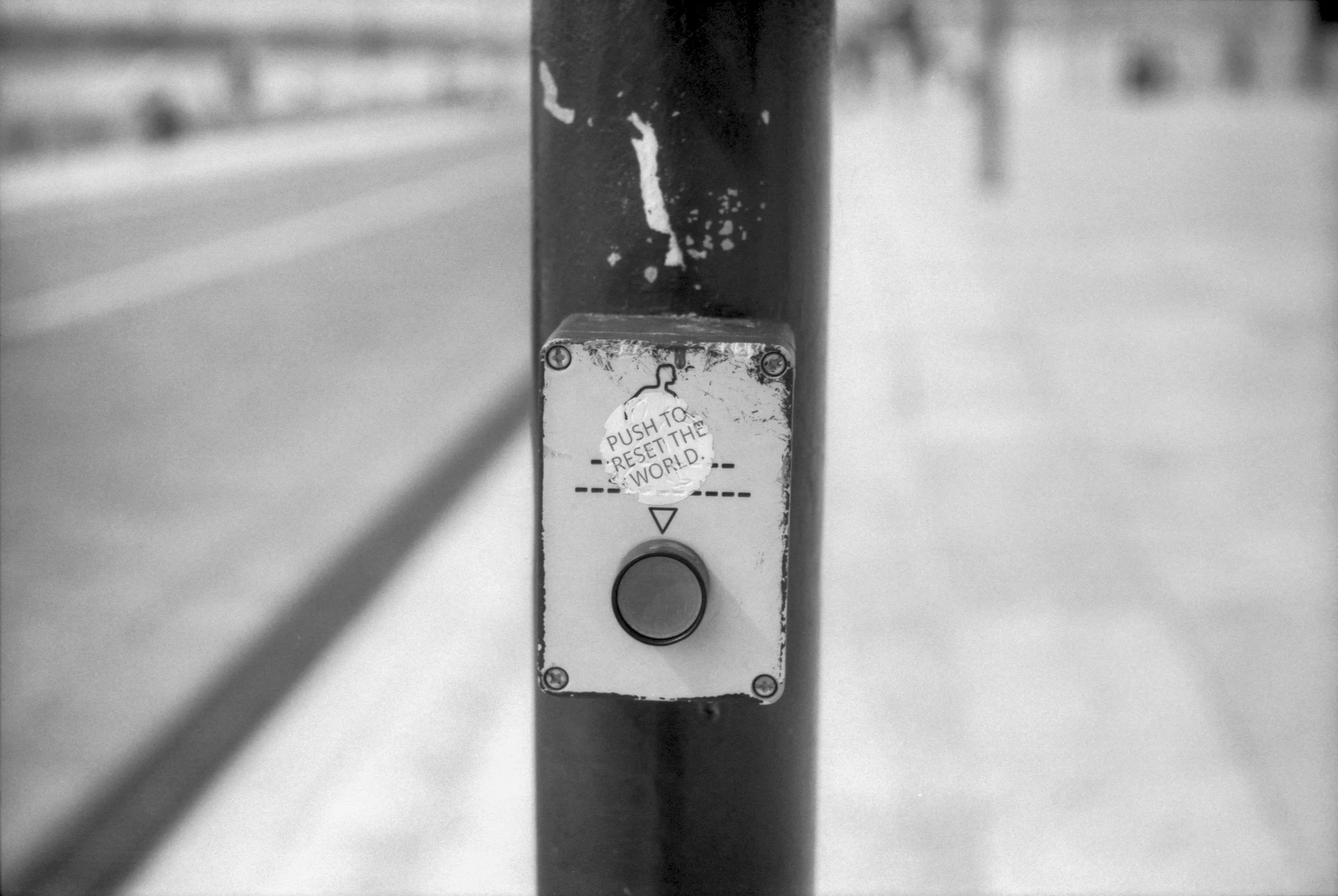 Close-up of a push button on a pole with a sticker above it that reads 'Push to reset the world' in black and white.