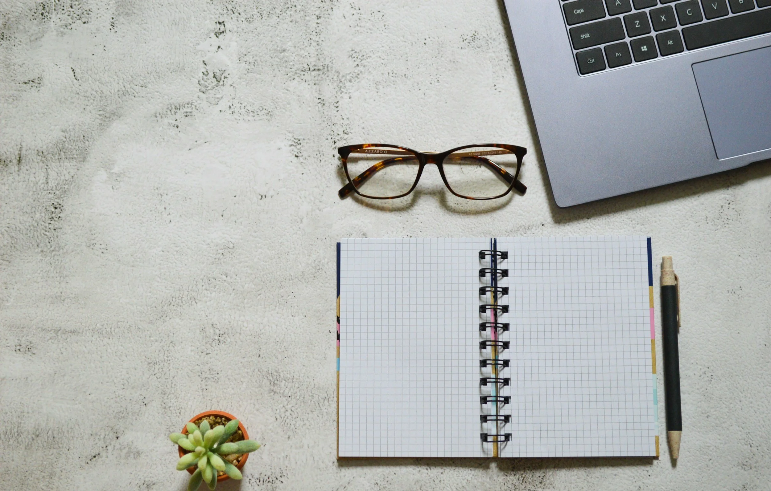 A flat lay photo showing a laptop, a pair of eyeglasses, an open graph paper notebook, a pen, and a small potted succulent plant on a textured white surface.