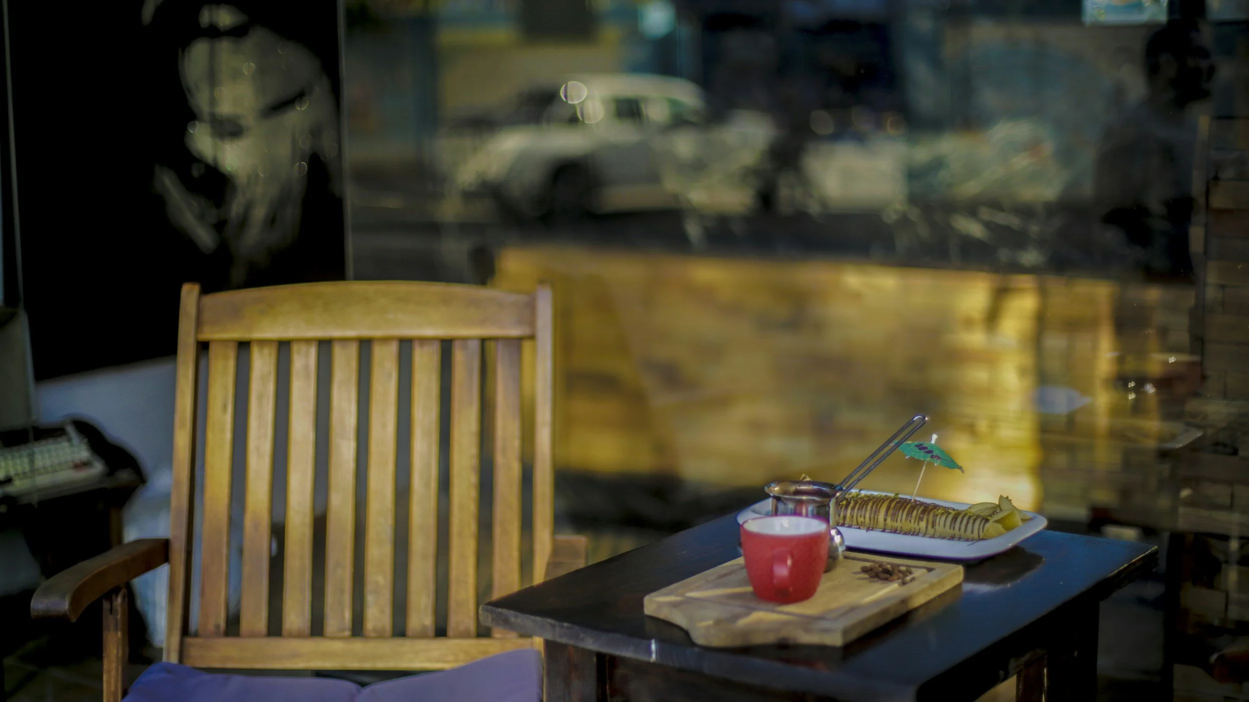 A cozy cafe setting with a wooden chair and a dark table. On the table, there is a wooden serving board with a red mug, a small metal container with a spoon, a plate with a rolled pastry garnished with a small umbrella, and some beans. The background shows a large window reflecting cars and a street scene outside.