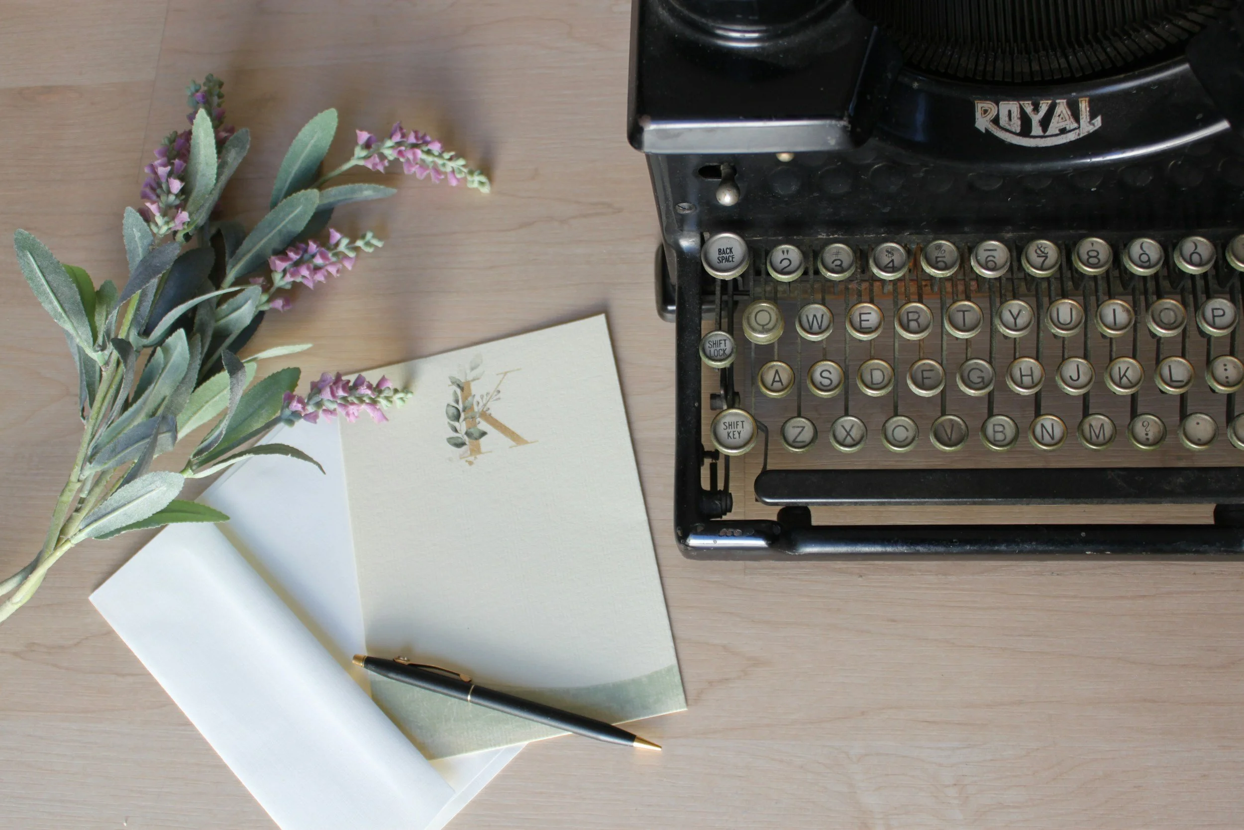 A vintage typewriter, a sprig of lavender flowers, an open notebook with a pen resting on it, and an envelope on a wooden desk.