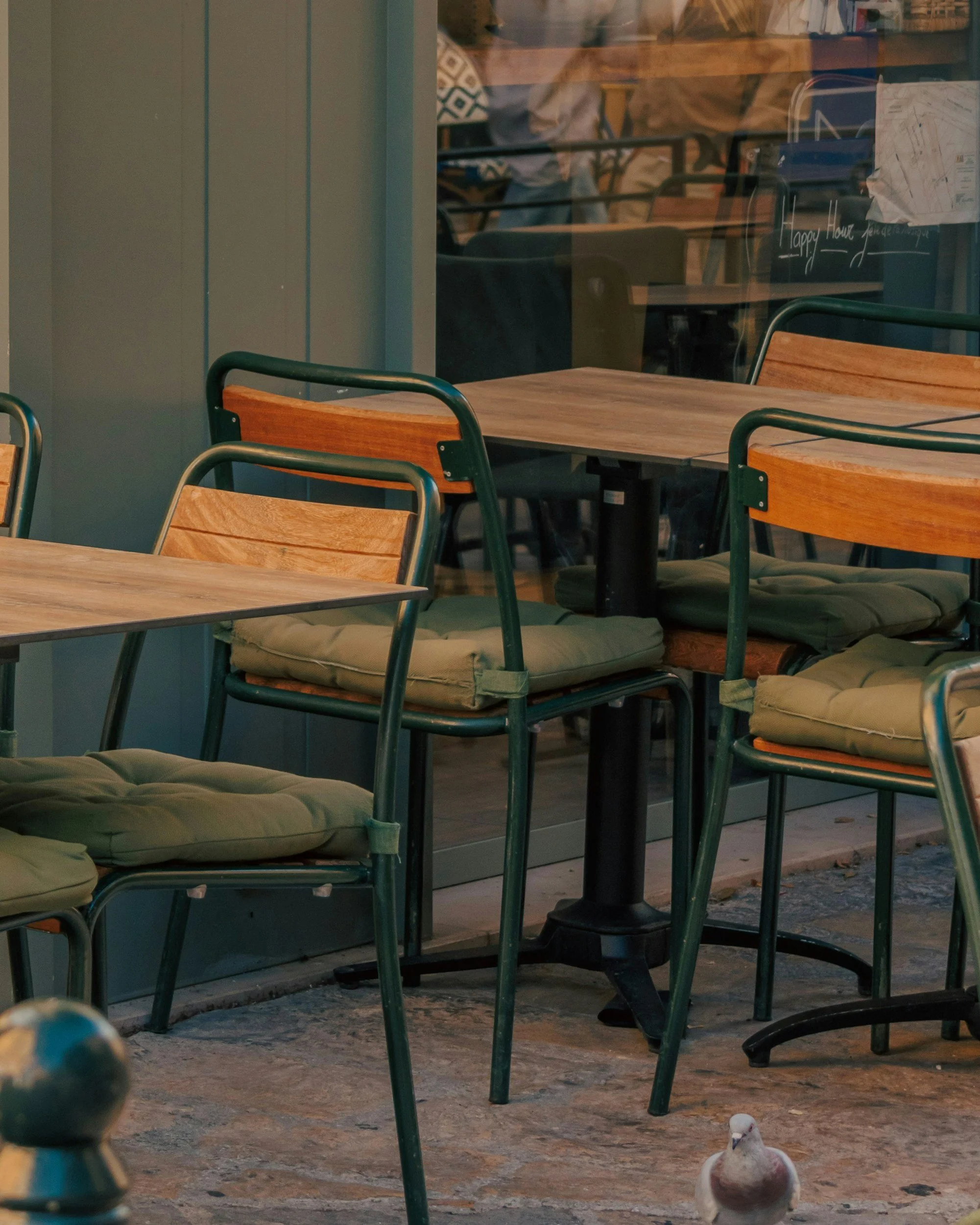 Empty outdoor cafe seating area with wooden chairs and beige cushions, viewed through a glass window with some reflections visible.