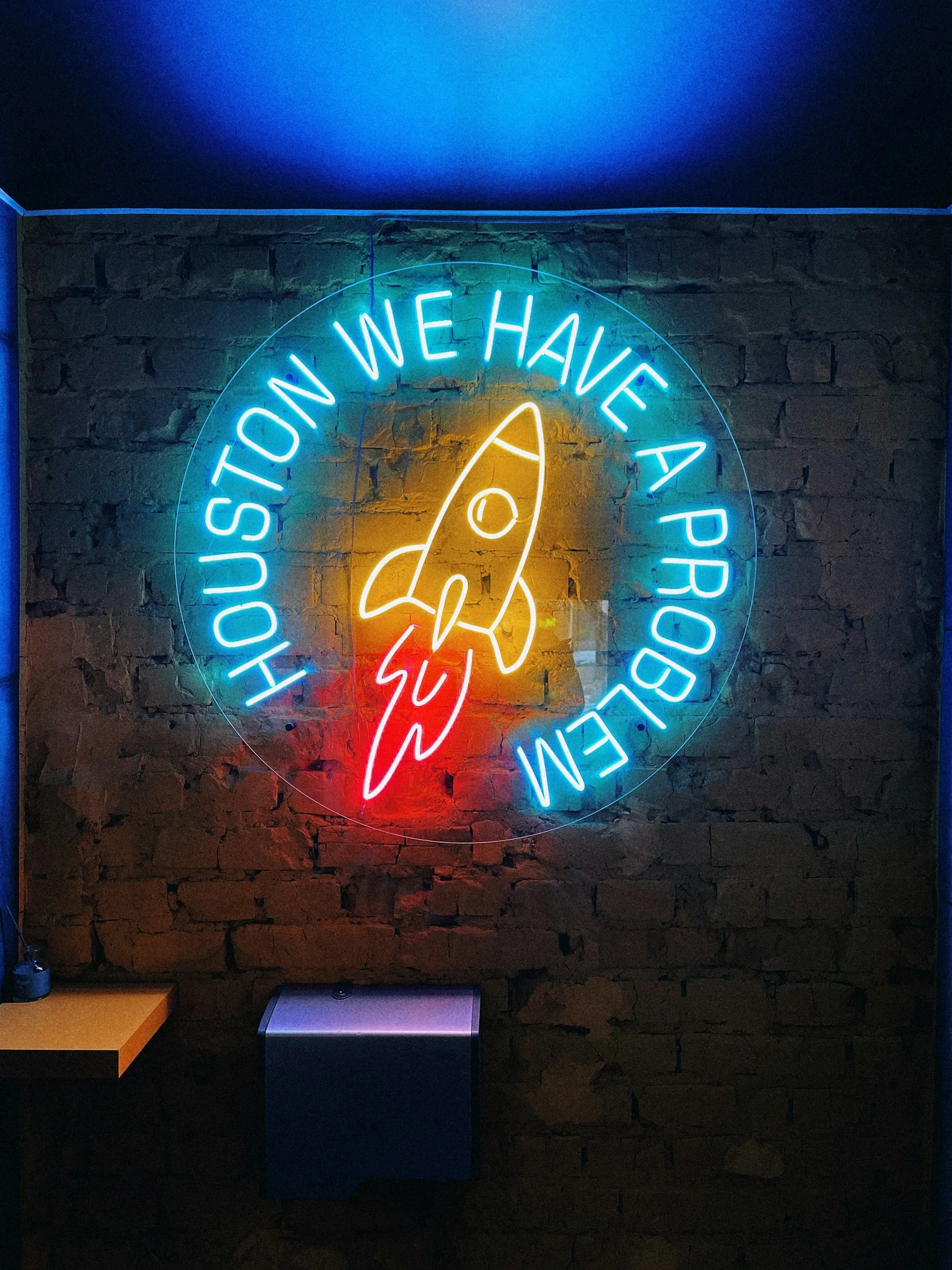Neon sign on brick wall with the text 'Houston We Have A Problem' and a rocket with a lightning bolt.