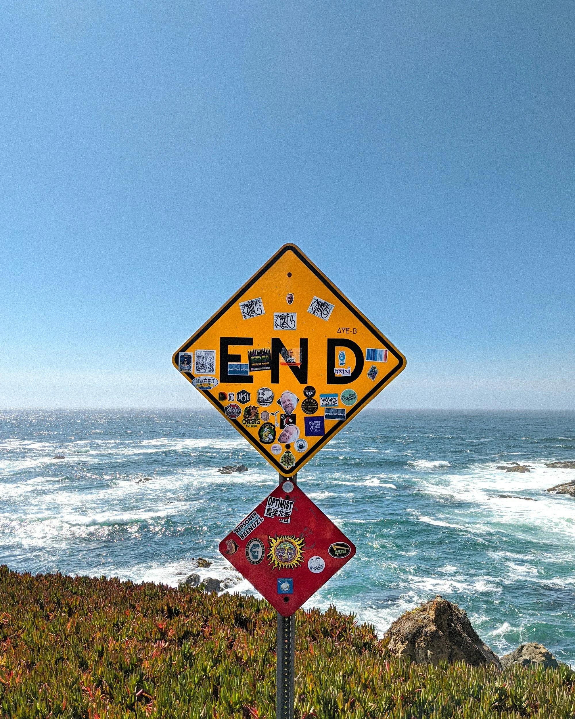 A yellow road sign with the word 'END' covered with various stickers, located by a rocky shoreline with ocean waves and a clear blue sky.