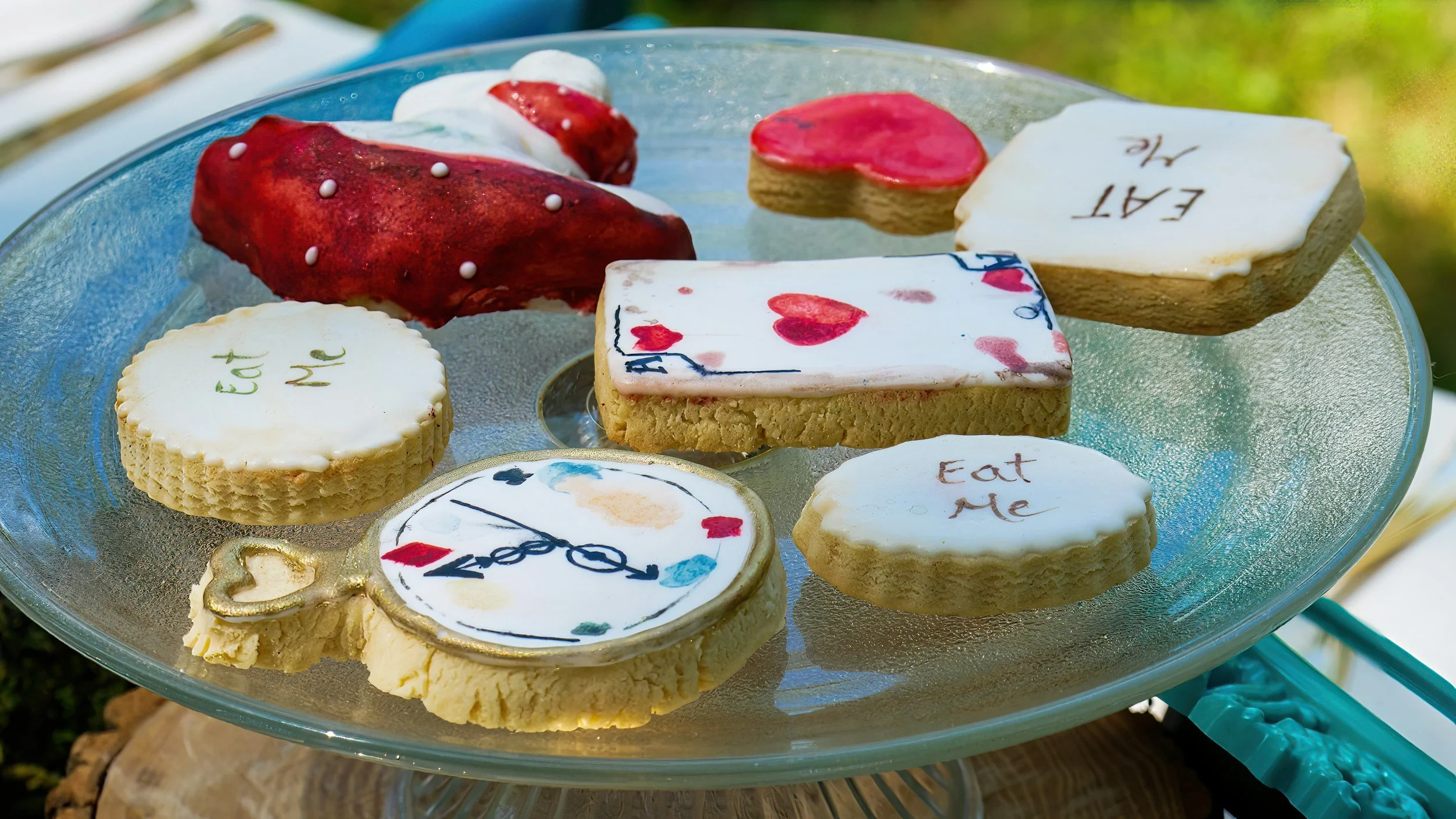 Assorted decorated sugar cookies on a glass plate, some with messages like 'Eat Me', 'Eat N', and others with designs like hearts, clocks, and strawberries.