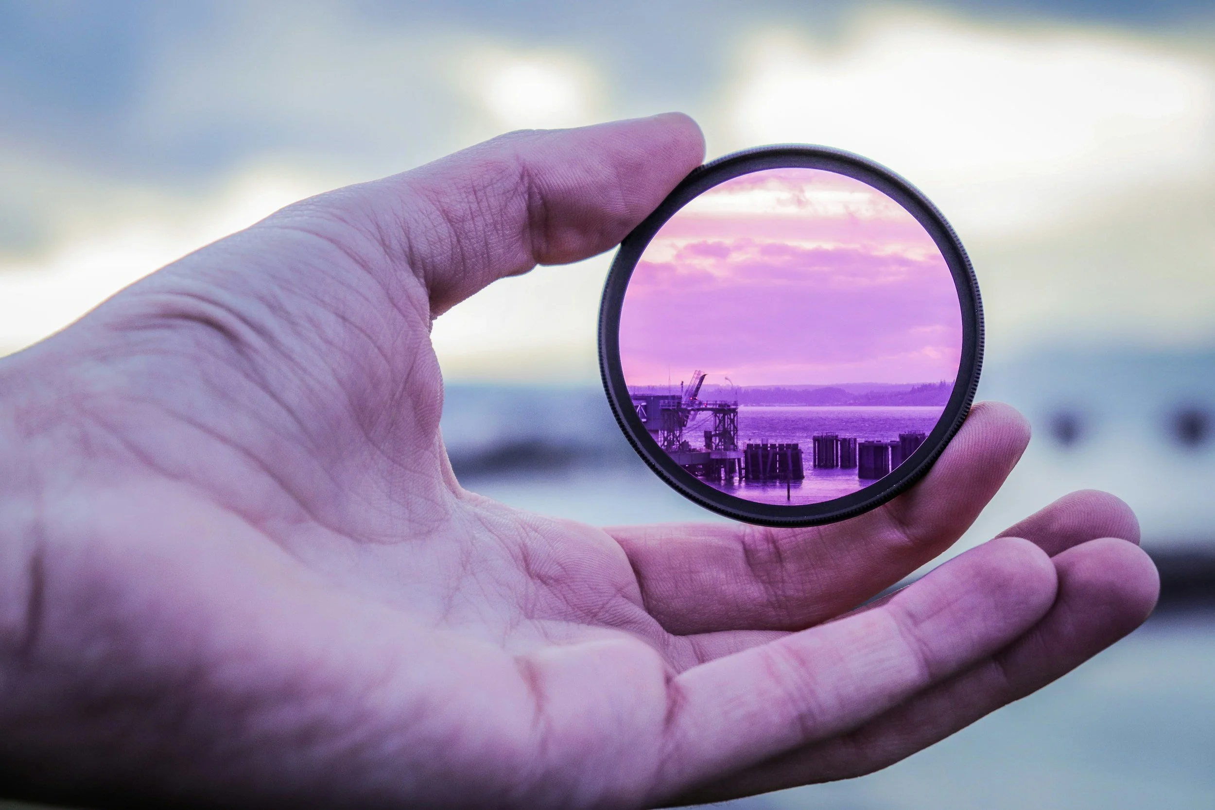 A hand holding a circular camera lens filter, which shows a pinkish-purple sky with clouds, a body of water, and an industrial dock with cranes and structures.