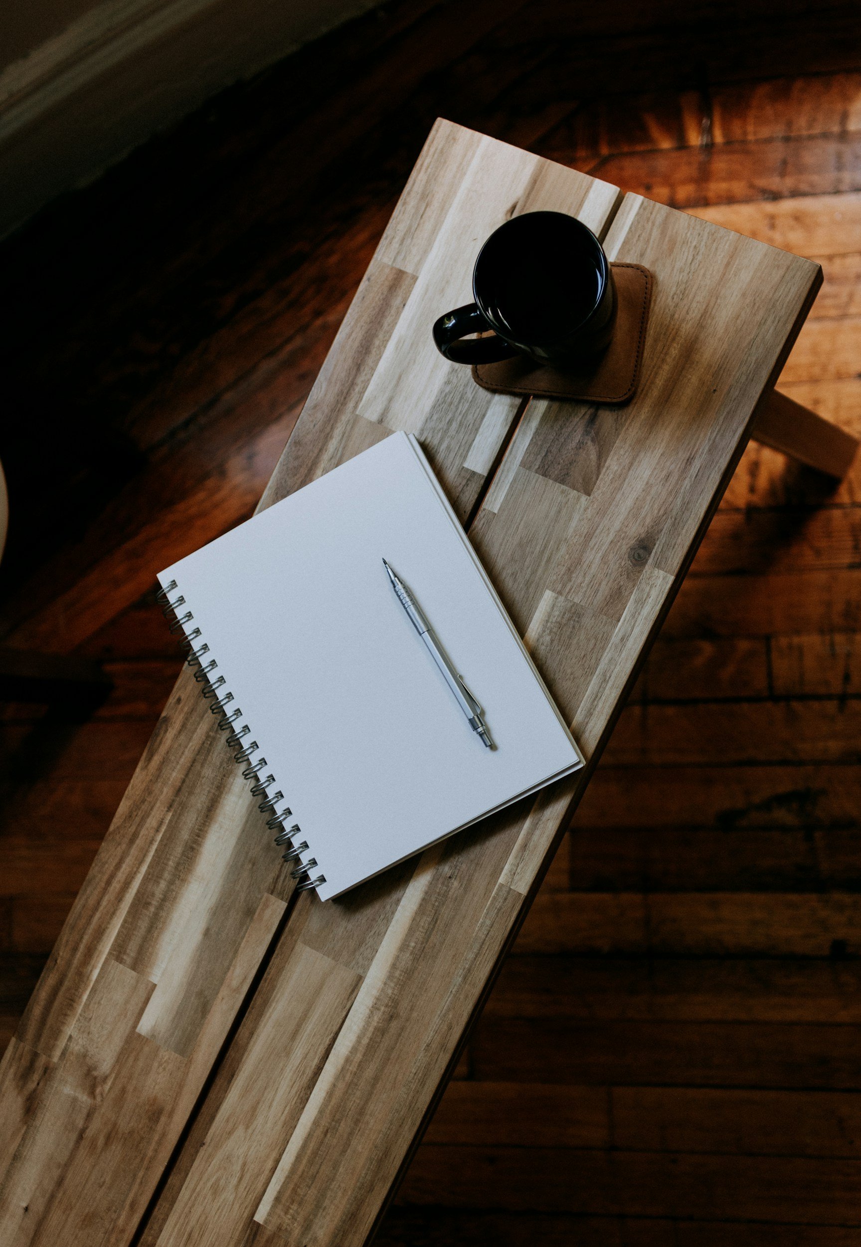 Top-down view of a wooden table holding a blank spiral notebook, a silver pen, a black coffee mug, and a brown coaster on a wooden table in a cozy room with wooden flooring.