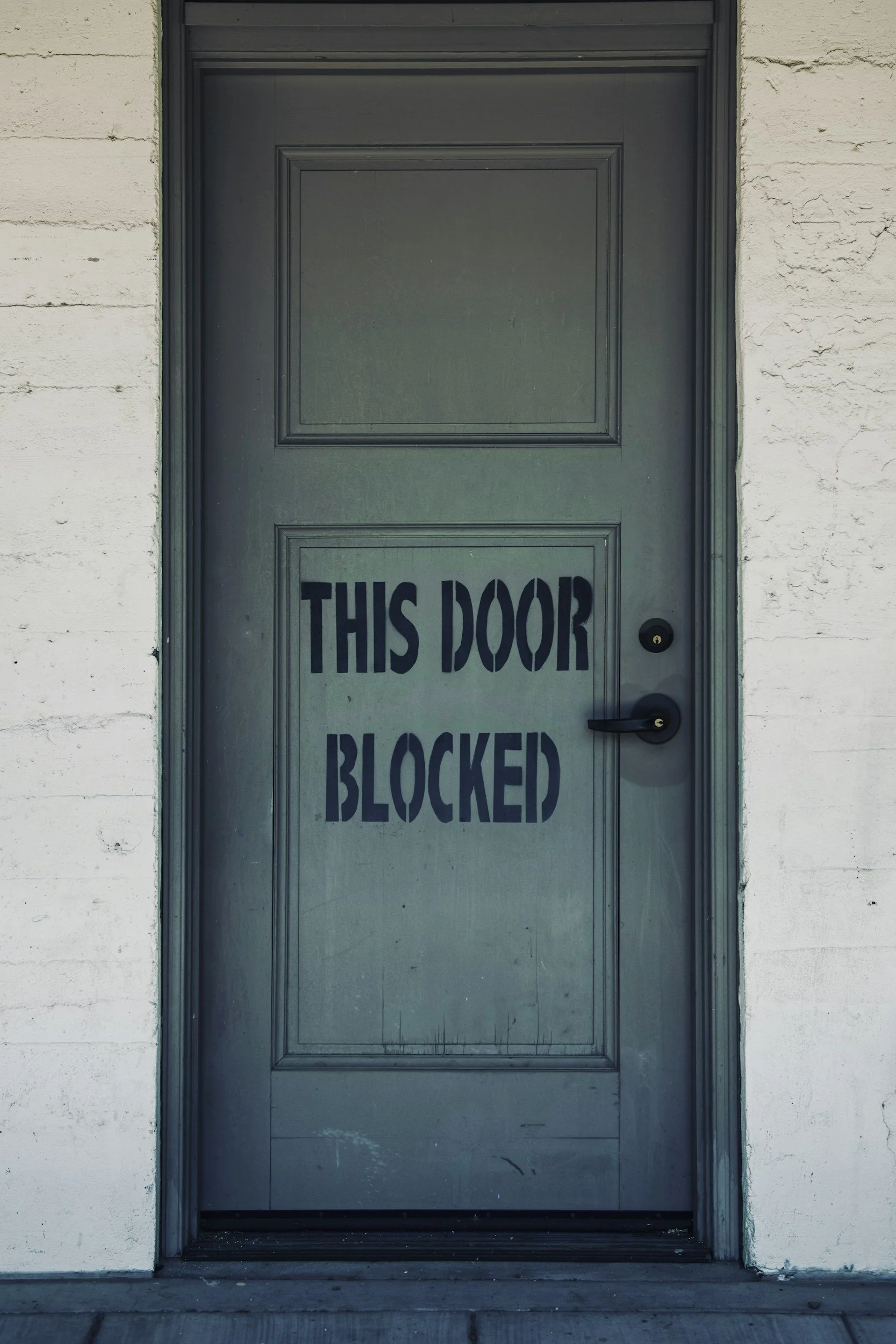 A gray door with a sign reading "THIS DOOR BLOCKED" painted on it, surrounded by white brick walls.