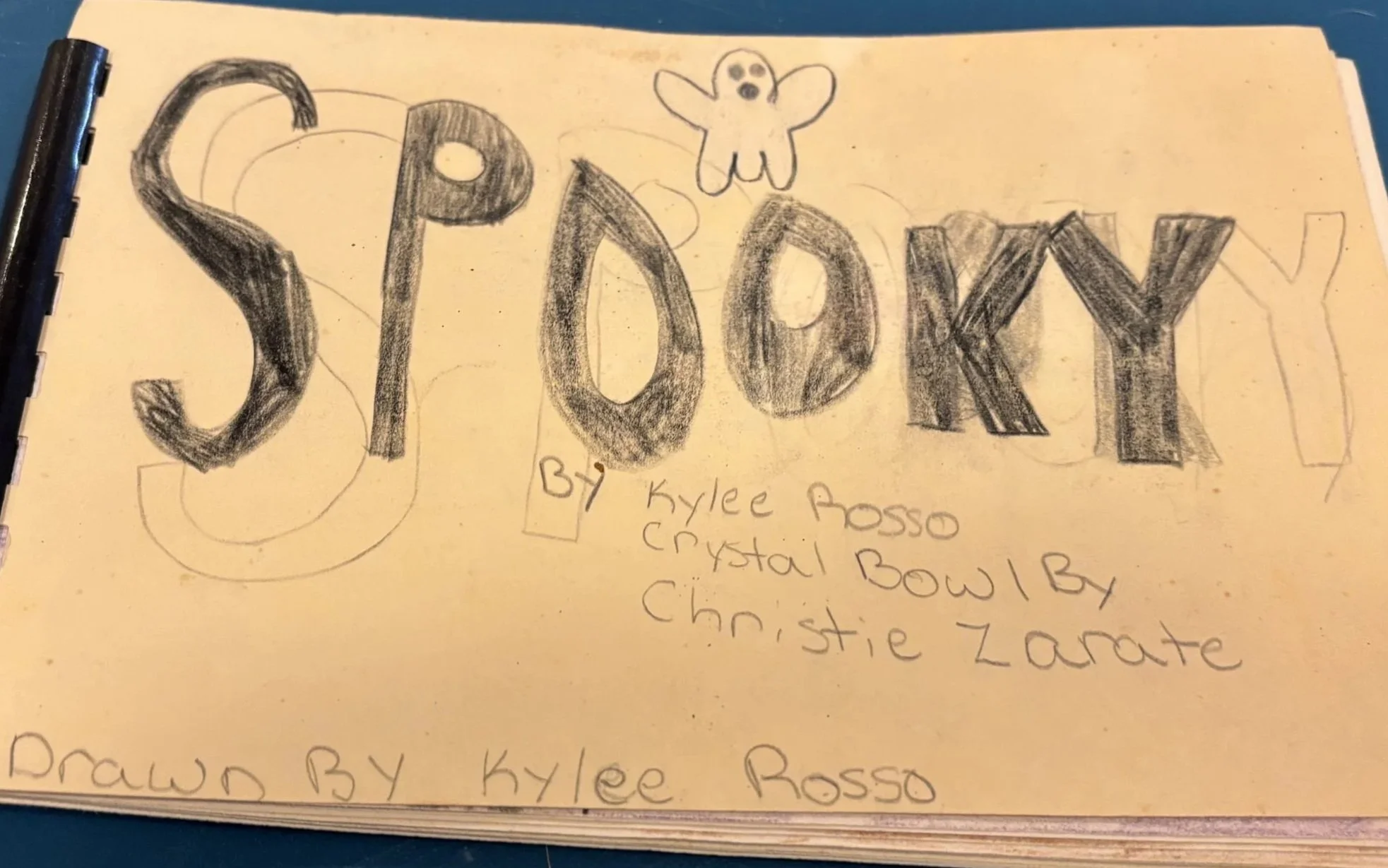 A hand-drawn title 'SPOOKY' with large, shaded letters and a small cartoon ghost at the top, written on yellow paper. Additional handwritten notes about the artist and contributors underneath.