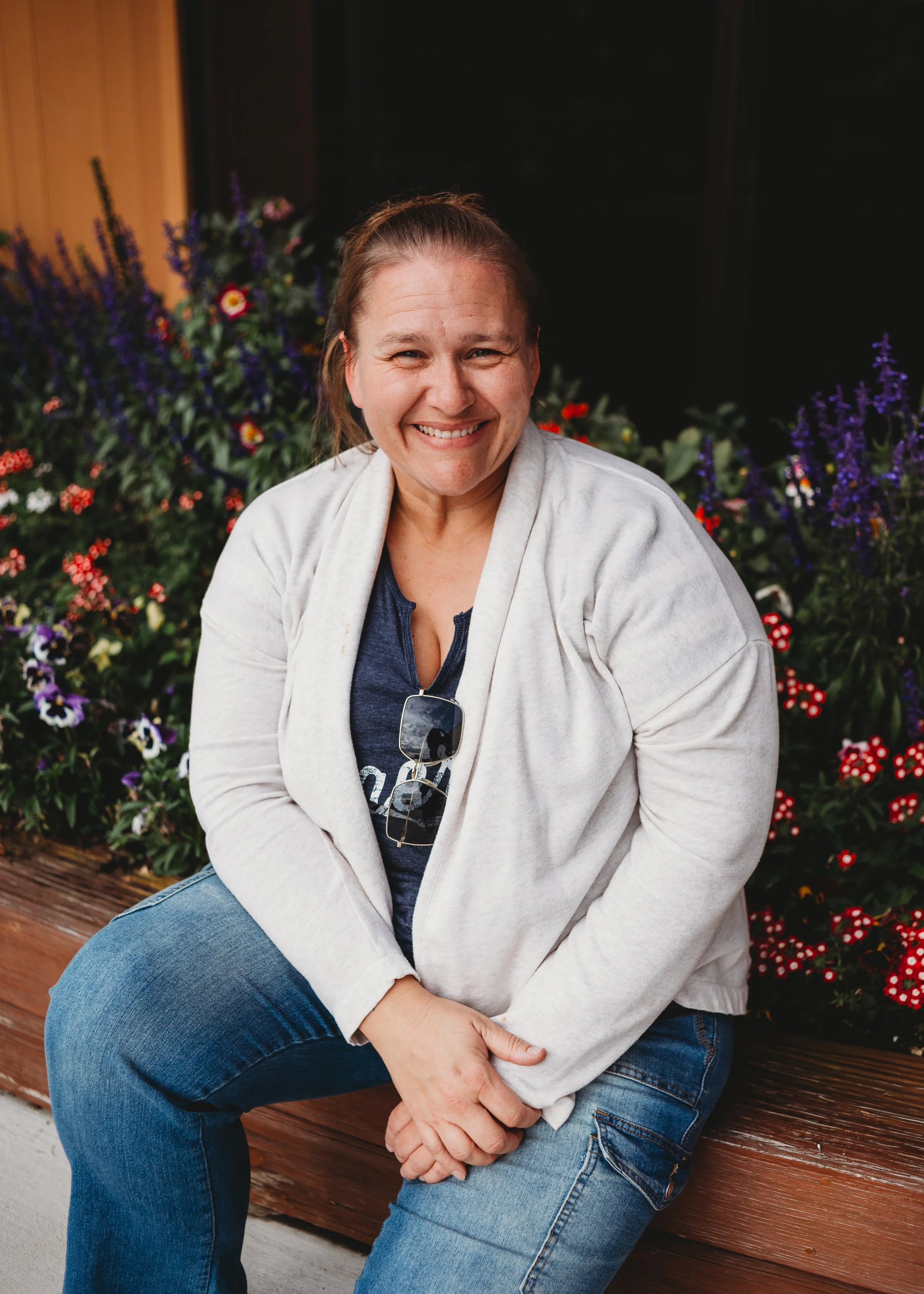 A woman smiling while sitting on a wooden bench with colorful flowers in the background.