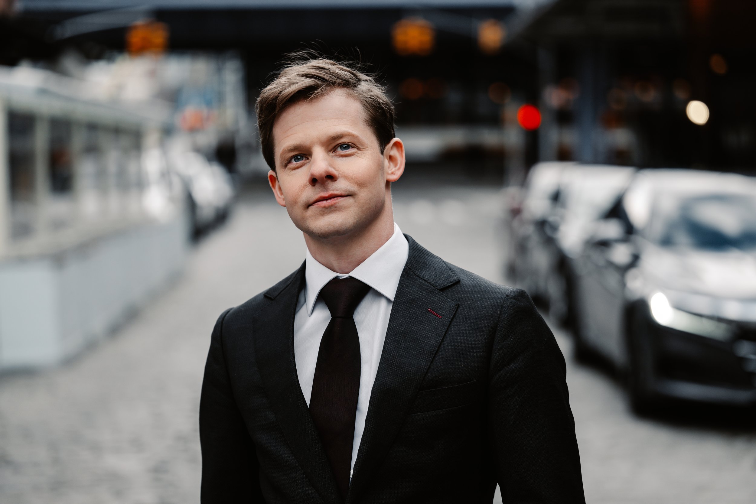 Mark Shuldiner in a black suit and tie standing outdoors in New York City with parked cars and a bridge in the background.