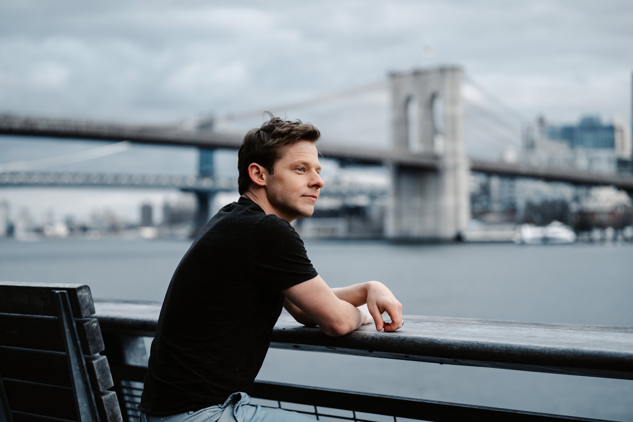 Mark Shuldiner sitting on a bench by the water, gazing pensively, with the Brooklyn Bridge in the background.