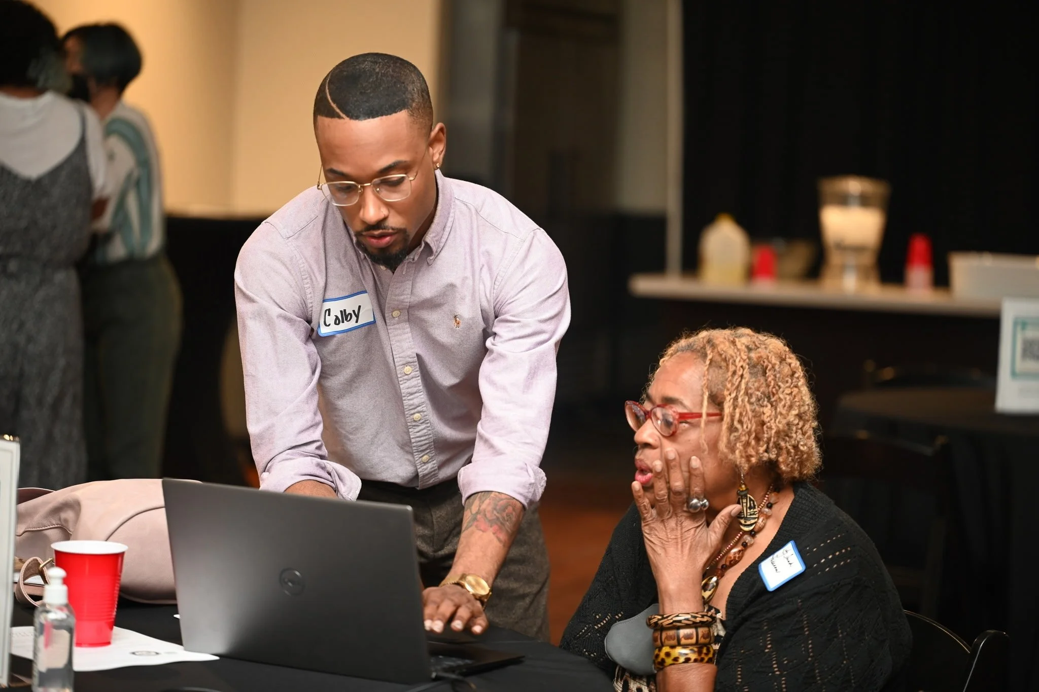 A young man with glasses and a name tag labeled 'Calby' is showing something on a laptop to an older woman with blond, curly hair and red glasses, who is reacting with surprise or amazement. They are at a table in an indoor setting.