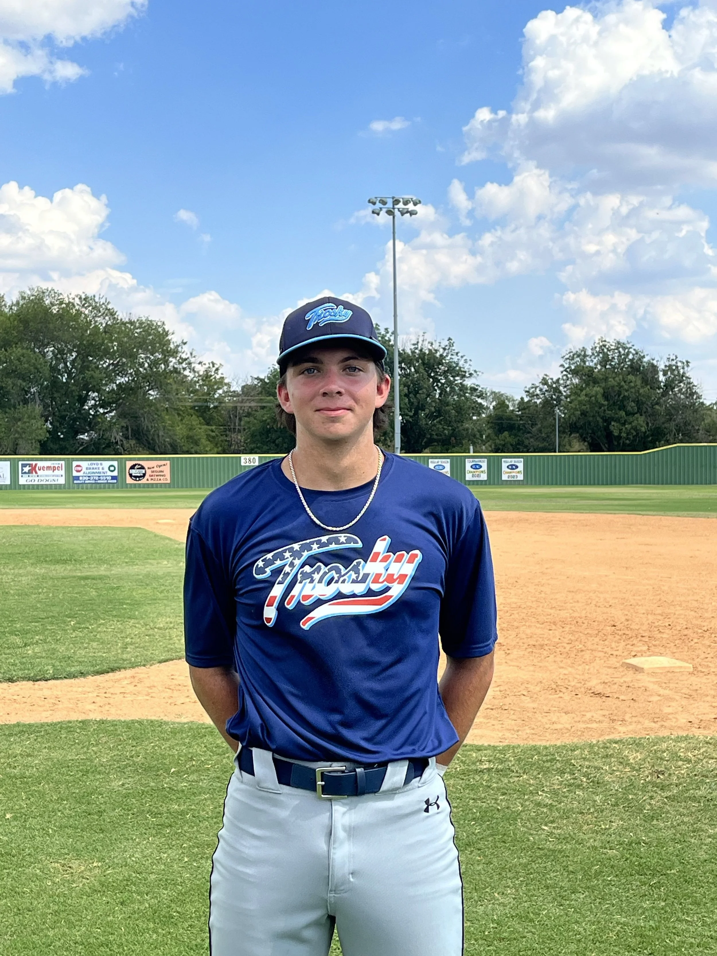 Young male baseball player in blue uniform standing on a baseball field with green grass, dirt infield, and trees in the background under a partly cloudy sky.
