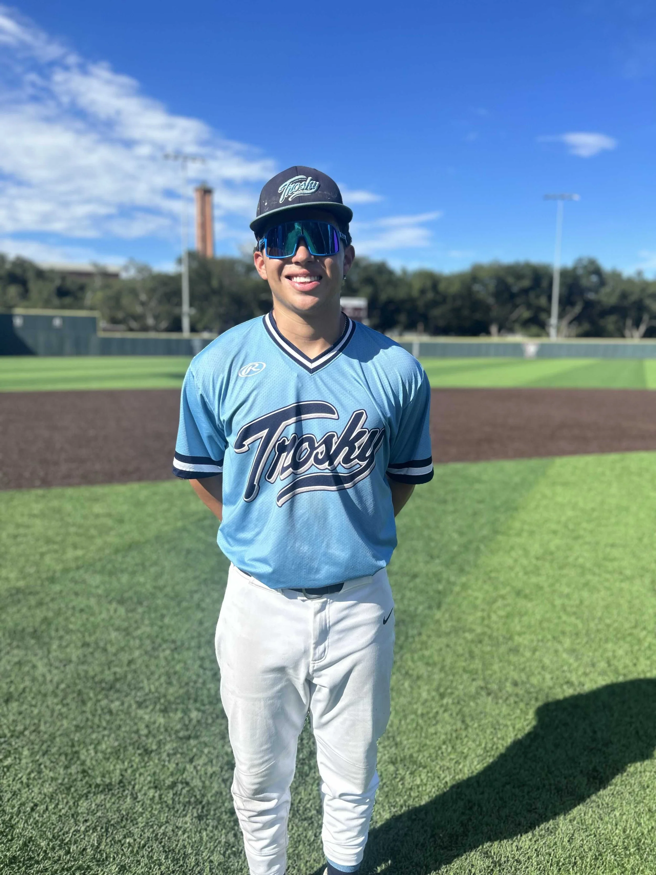Young male baseball player in a blue jersey and white pants, wearing sunglasses and a cap, standing on a baseball field on a sunny day.