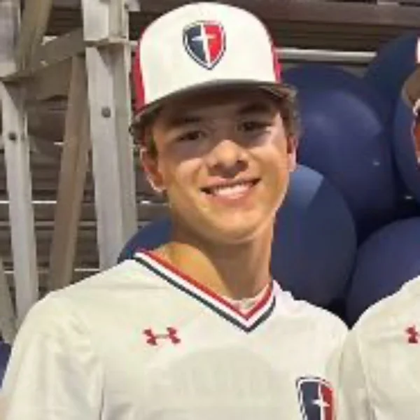 Young man wearing a white sports jersey and a baseball cap with a letter T logo, smiling at a sports event.