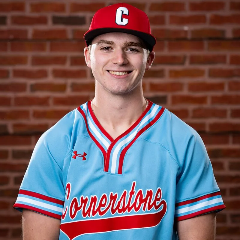 A young man wearing a red baseball cap with a white letter C, a light blue sports jersey with red and white stripes, and 'Conerstone' written across the front, smiling in front of a brick wall.