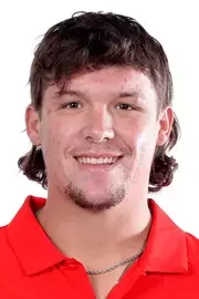 Close-up of a young man with short dark hair, wearing a red polo shirt and a necklace, smiling against a plain background.