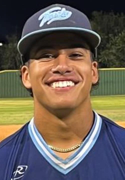 Young man smiling, wearing a baseball cap, and baseball jersey on a baseball field at night.
