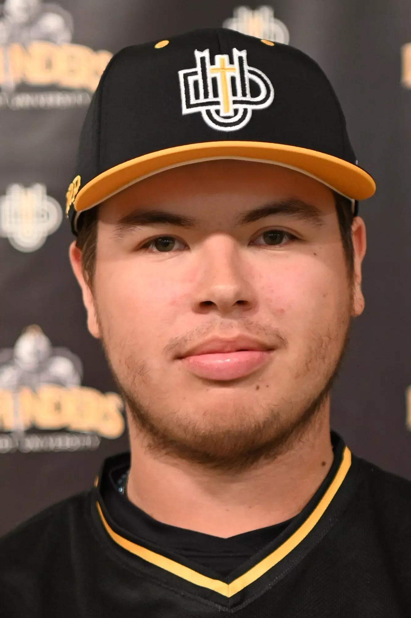 A young man wearing a black baseball cap with yellow accents, featuring a logo with a cross and initials, and a black sports jersey with yellow trim.