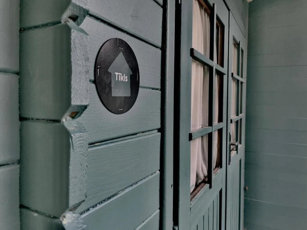 A close-up of a wooden exterior wall with a black circular sign featuring an upward arrow and the words 'Tikis' on it. To the right, there is a set of wooden double doors with glass panes and curtains behind the glass.