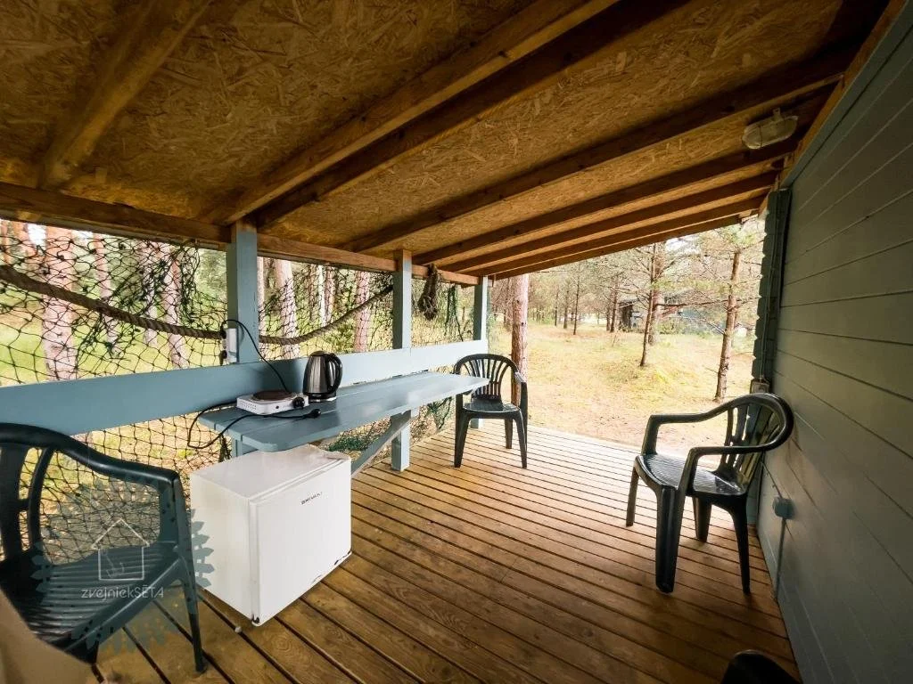 Covered wooden porch with three black plastic chairs, a white mini fridge, a small black electric stove, a kettle, and a white wall-mounted light fixture, overlooking a wooded outdoor area.