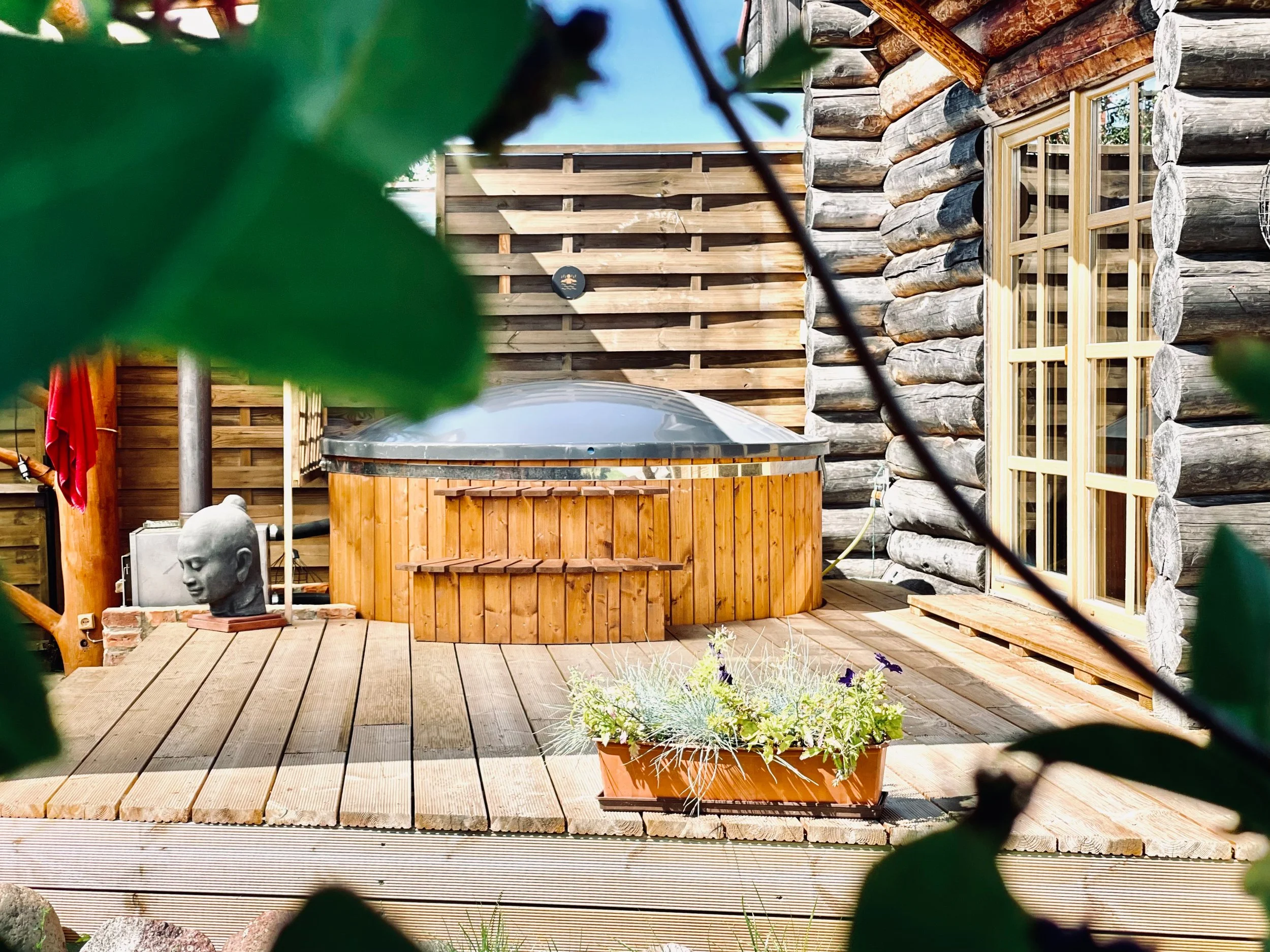 A wooden deck with potted plants, a hot tub surrounded by a wooden exterior, a sculpture of a human head, and a log cabin-style house with large windows, during daytime.