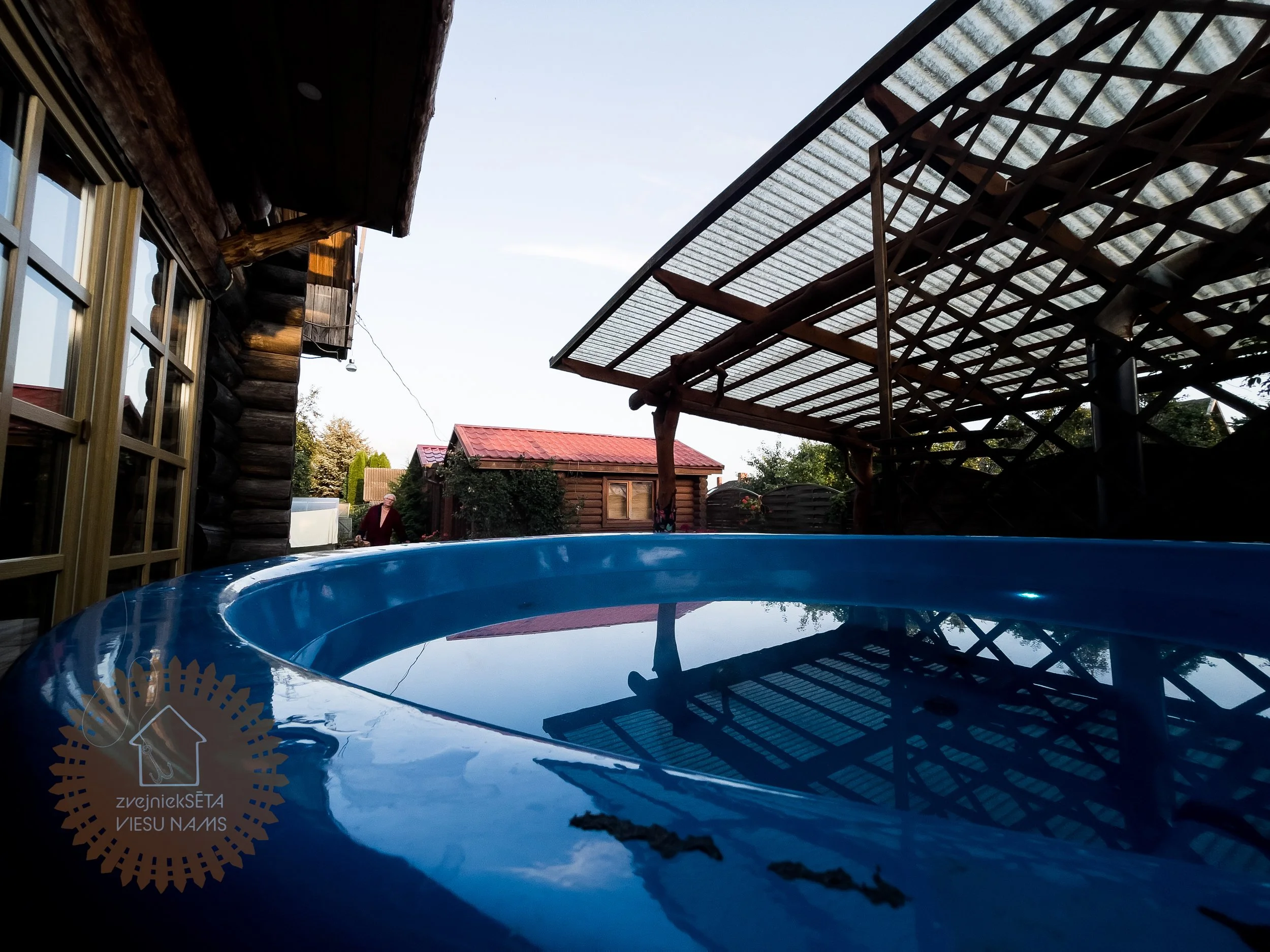View of a wooden cabin's side with a window, an outdoor wooden structure with a corrugated metal roof, and a blue plastic pool filled with water in a backyard yard with trees and a red-roofed shed.