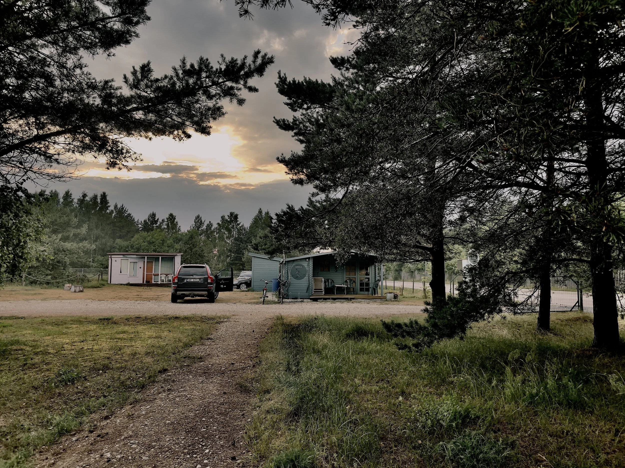 A rural campsite at sunset with trees framing small cabins, parked cars, and bicycles.
