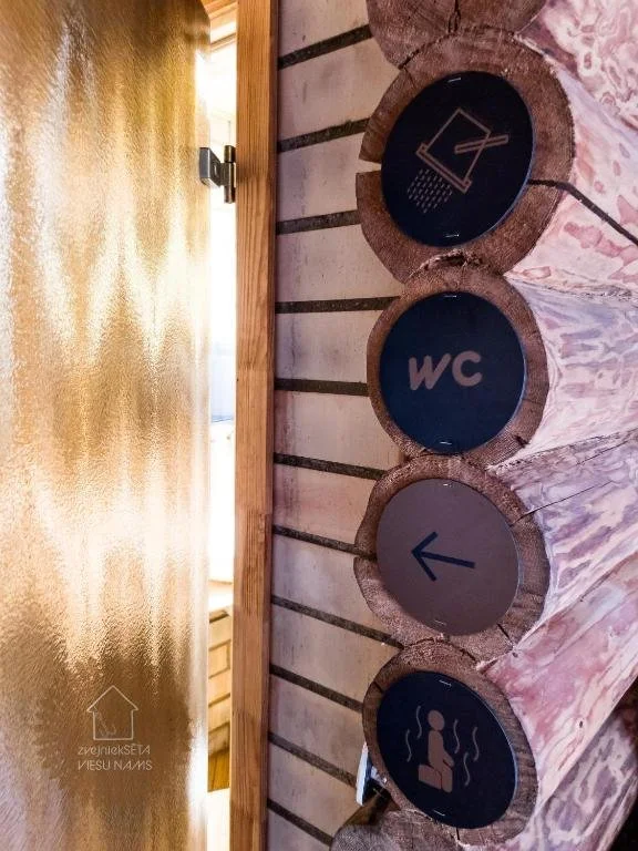 Vertical wooden wall with four round signs indicating a shower, a women's restroom, a left arrow, and a heating or hot water symbol next to a textured yellow wall and doorway.