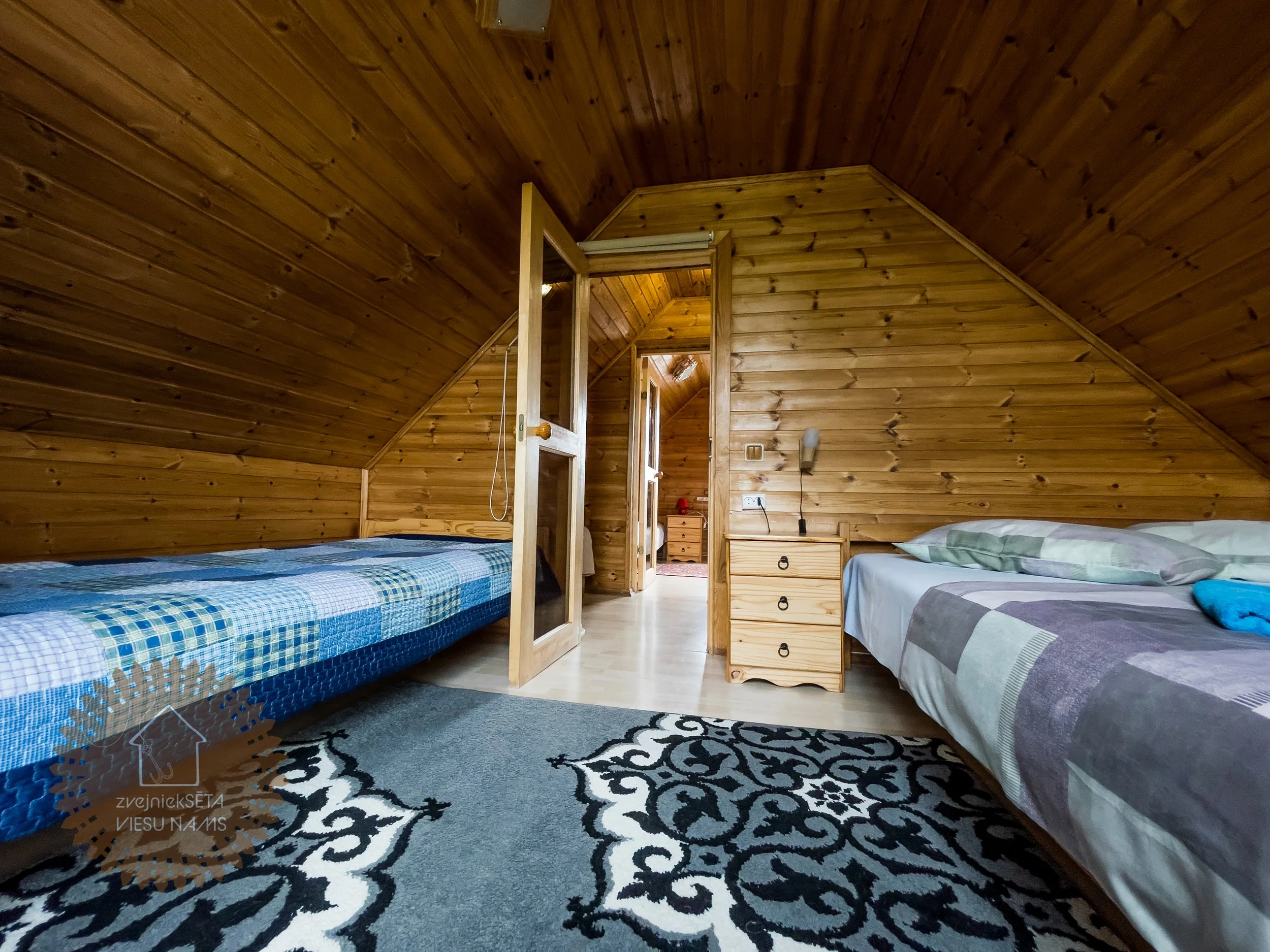Attic bedroom with wood-paneled walls and ceiling, two beds with patterned bedding, a small wooden nightstand with drawers, and a patterned rug.