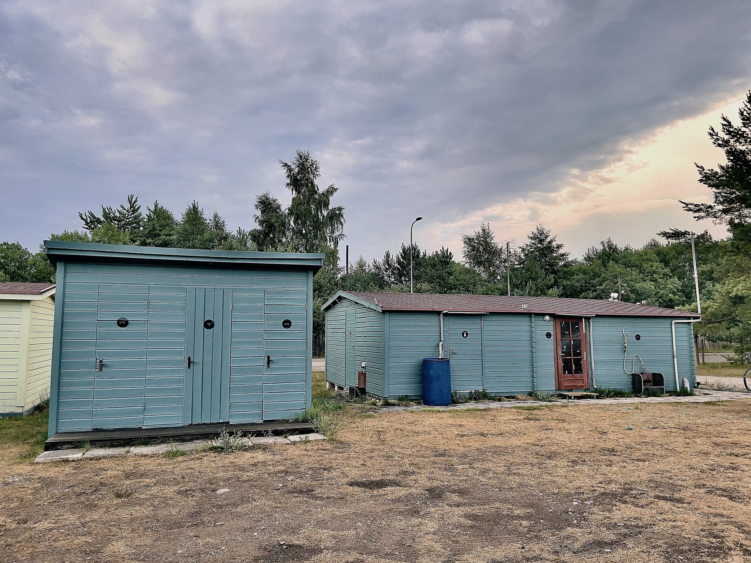 Two small, teal-colored sheds with metal siding and one with a brown door, one with a closed door, and a large blue barrel outside the larger shed. The ground is dirt with some sparse grass, and there are trees and sky with clouds in the background.