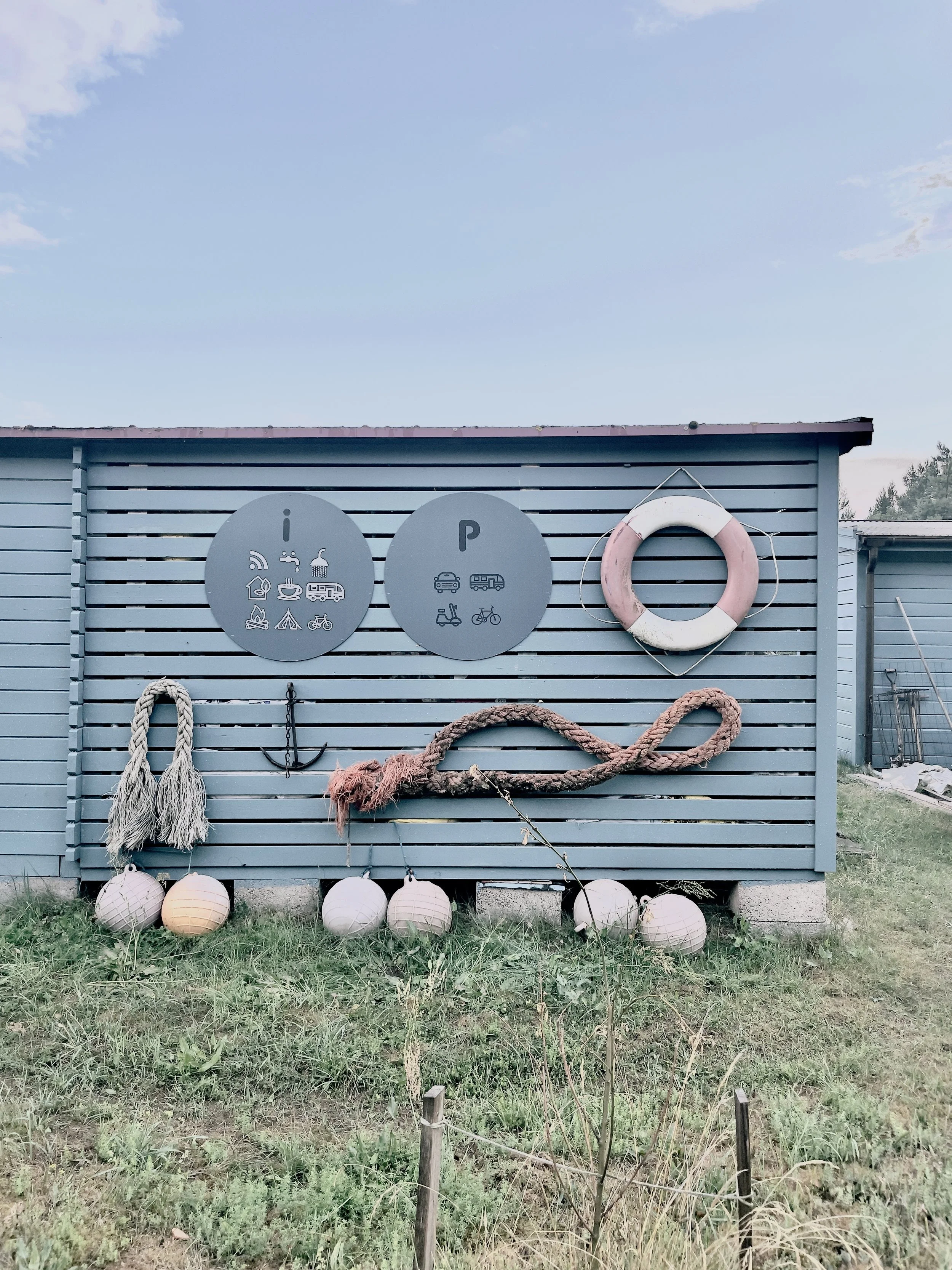 Decorative outdoor wall with nautical and camping theme, including life buoy, rope, and lanterns, set against a blue slatted fence with signage for camping and parking.