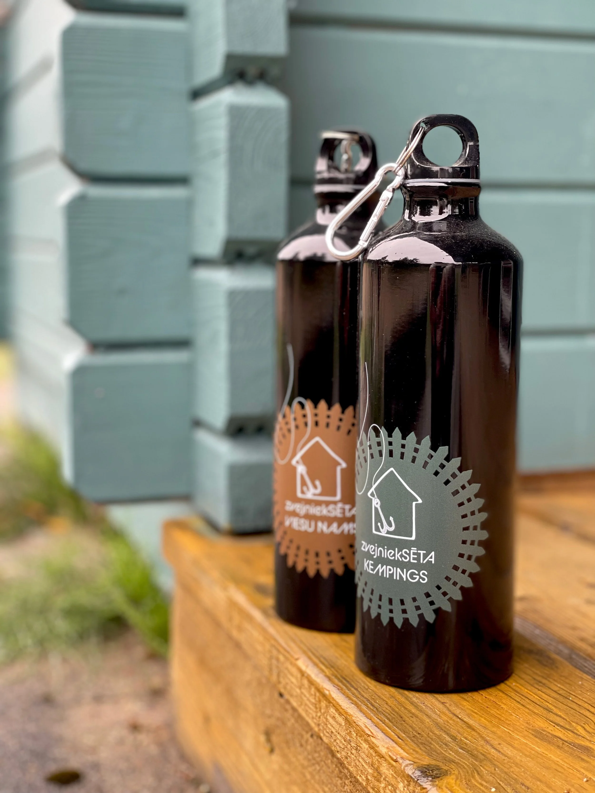 Two black bottles with white and orange labels featuring a house and tree icon, sitting on a wooden surface outdoors, with a background of stacked light gray containers or drawers.