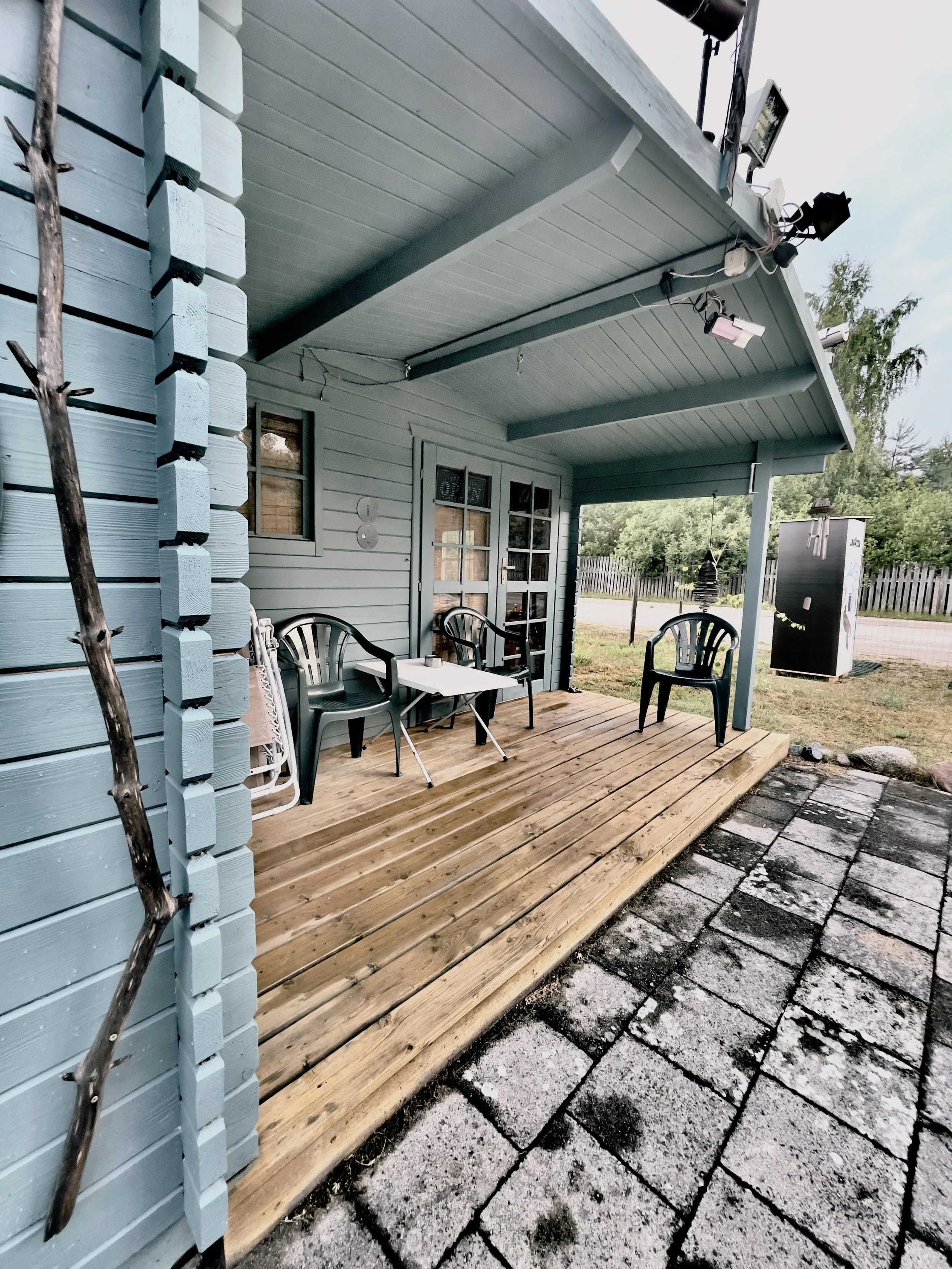 A patio with a wooden deck attached to a light blue house, featuring three plastic chairs, a small white table, and outdoor lighting fixtures on the ceiling. There is a window and a door with glass panels, and a black utility box outside, with a fenc
