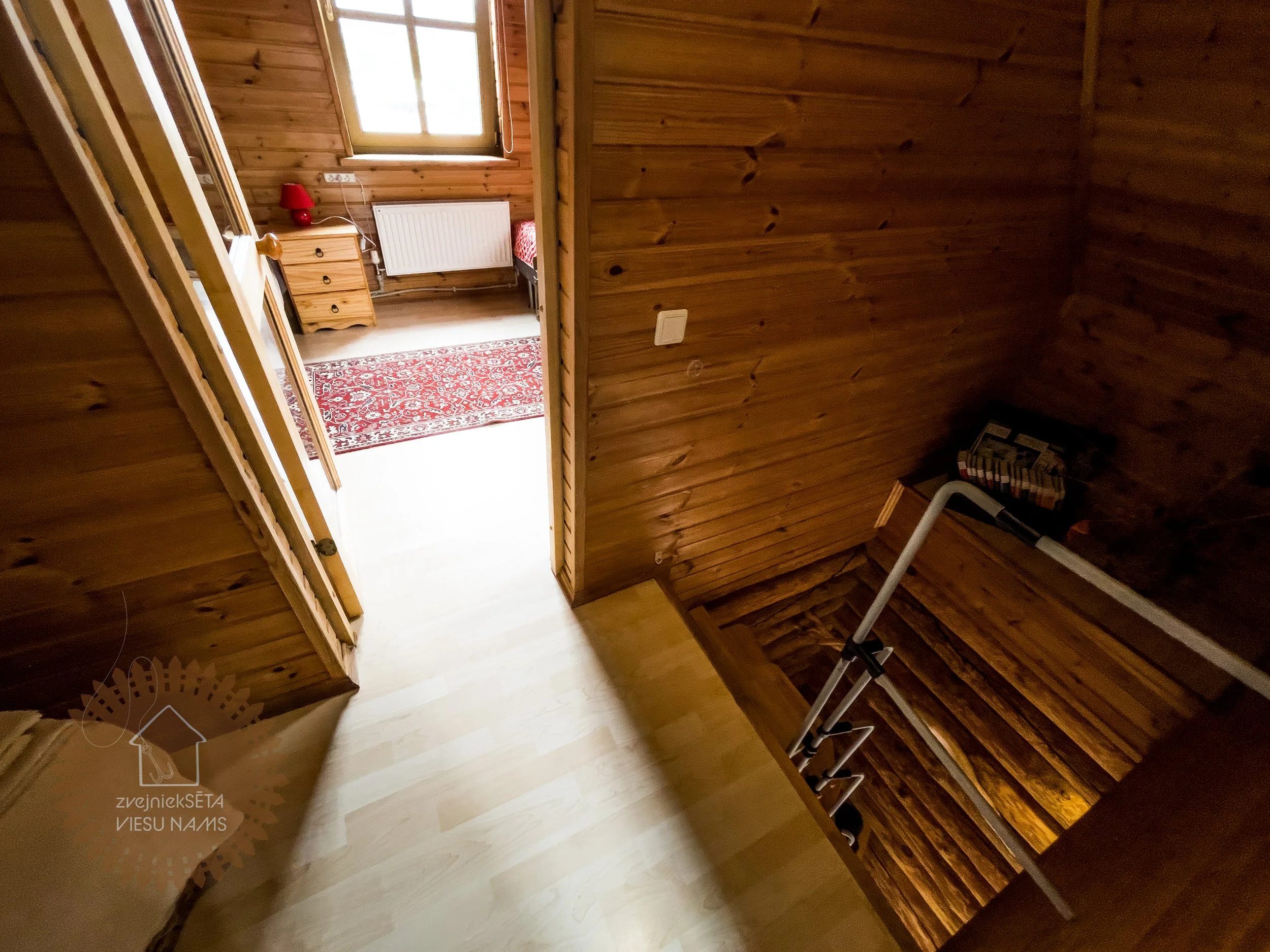 View of a wooden staircase leading to a second floor from the top, with a hallway and a bedroom visible at the end of the hall. The bedroom has wooden walls, a window, a nightstand with a lamp, a radiator, a bed with a pink blanket, and a red pattern
