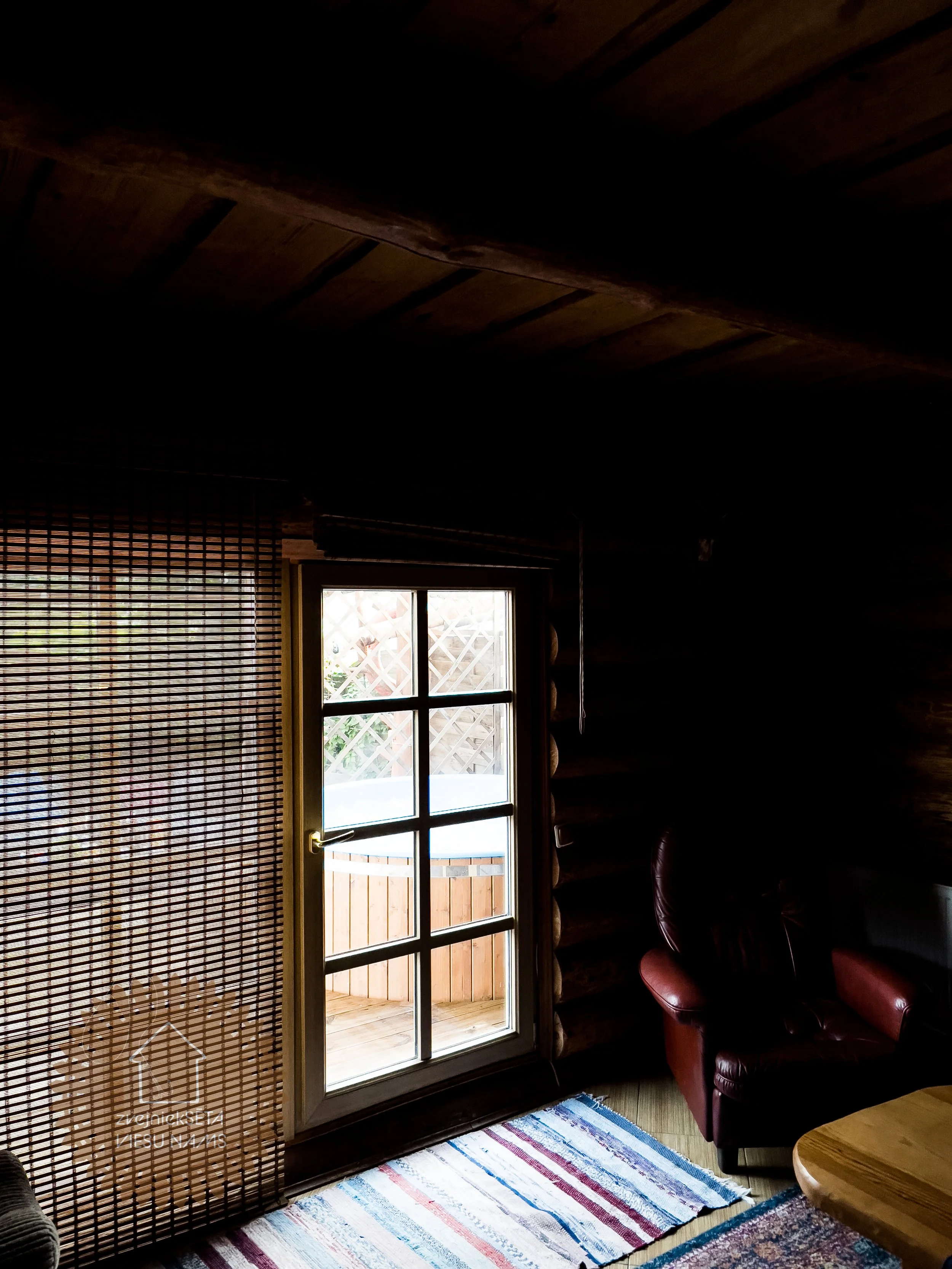 Interior of a cozy cabin with a glass door leading outside to a balcony, a woven curtain, a red leather armchair, and a multicolored rug on a wooden floor.
