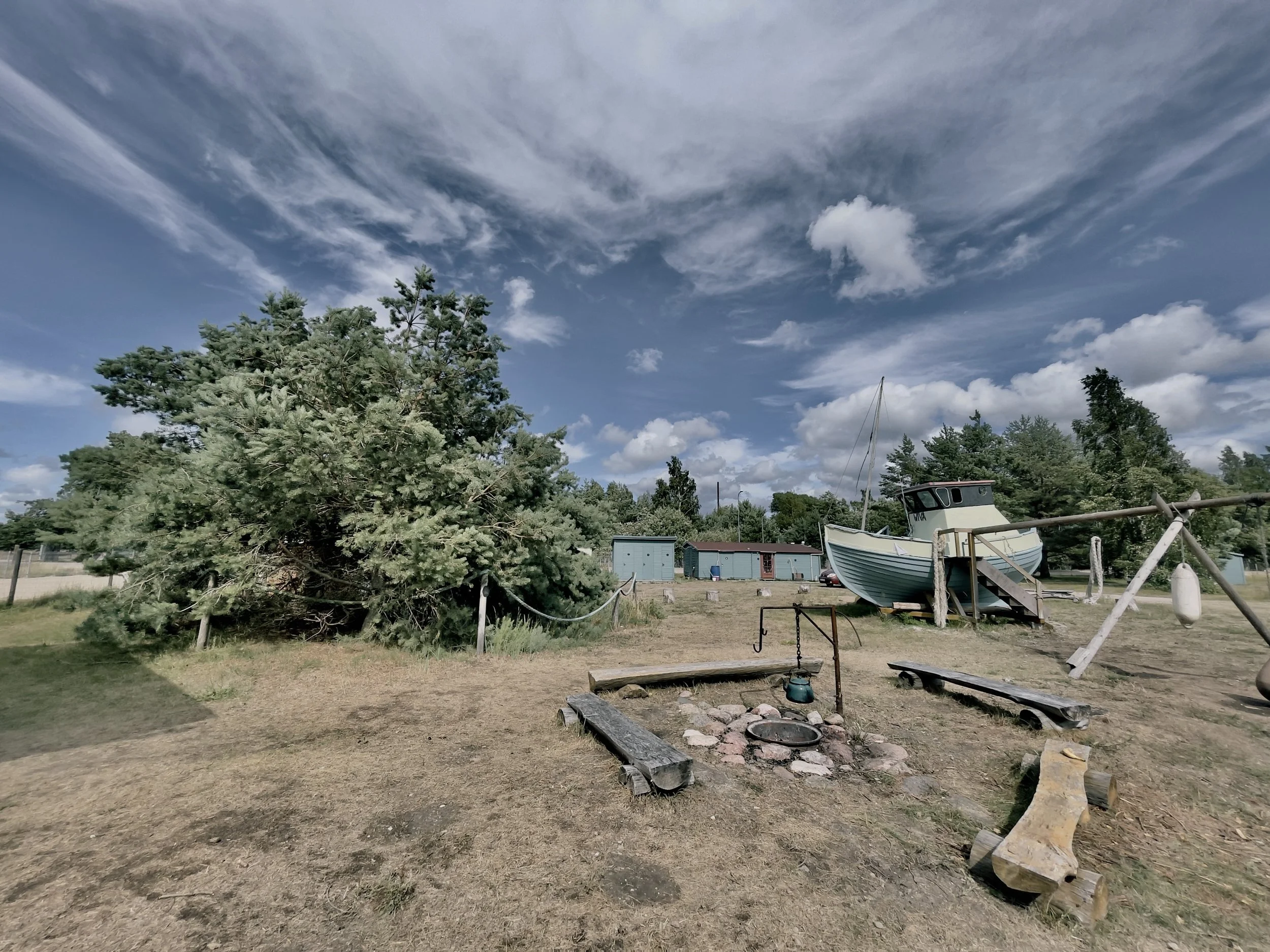 A playground with a small fire pit and benches, a large tree, a boat on a stand, some small storage sheds, and a swing set on a grassy area under a sky with clouds.