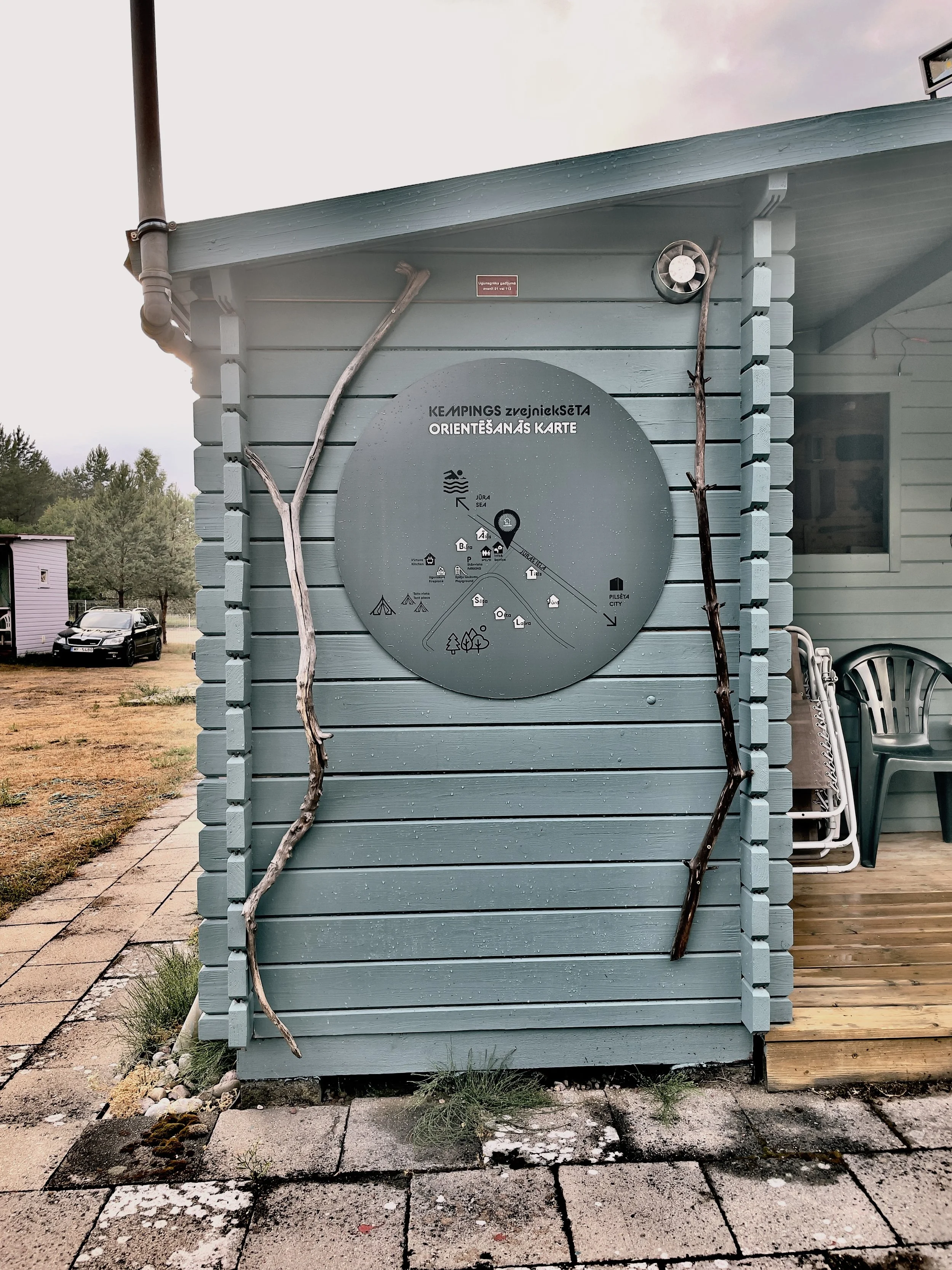 A wooden building painted light blue with a round map sign on the wall showing a camping and hiking trail, with two pieces of weathered sticks mounted vertically on either side of the sign, and a chair and some stacked white chairs on the porch to th