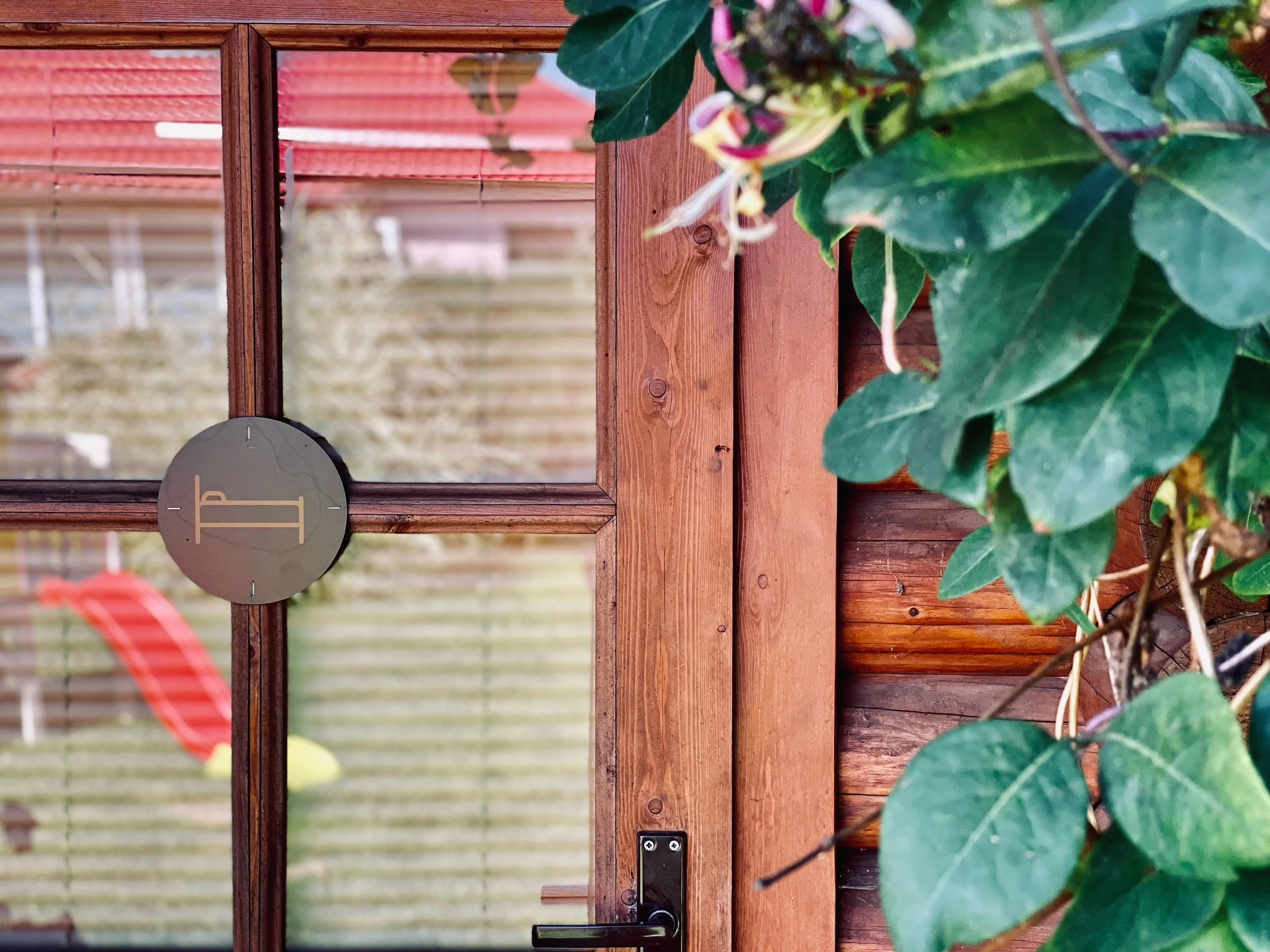 Close-up view of a wooden door with a black handle, a circular black and gold decorative piece, and green leafy plants on the side.